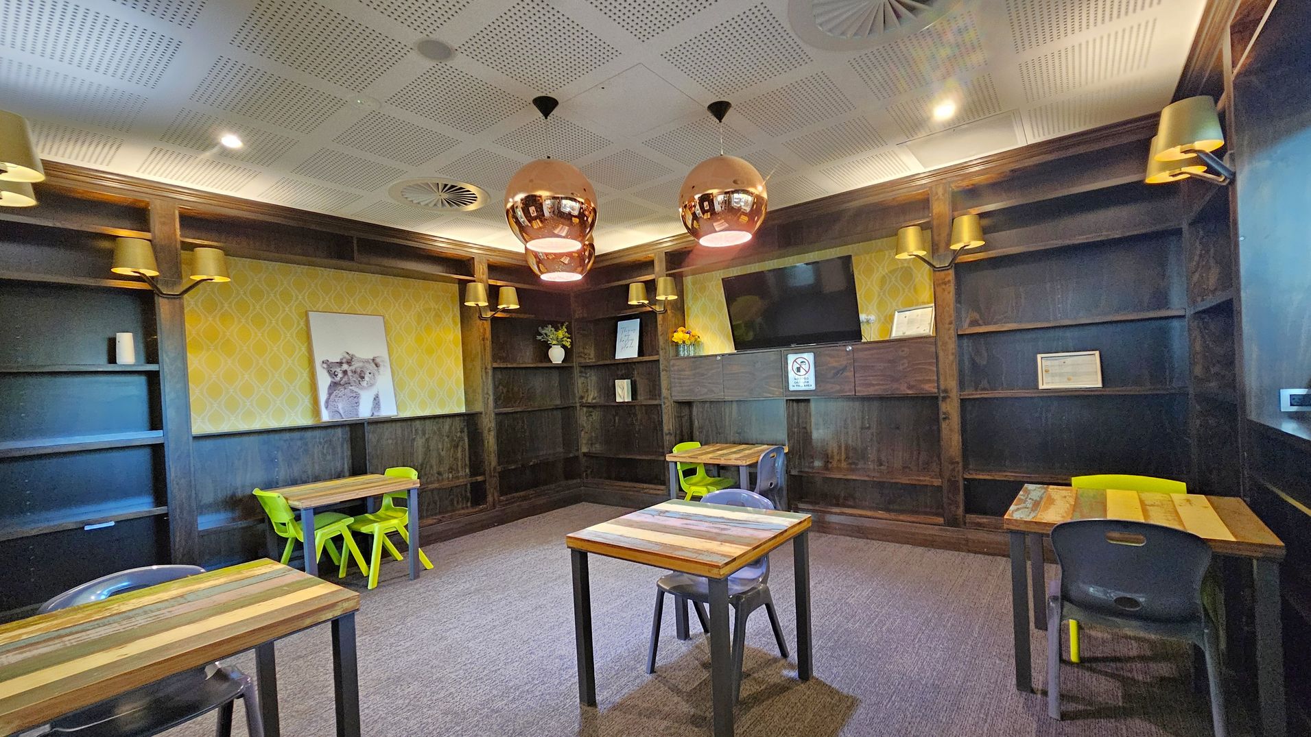 A room with dark wooden shelves lining the walls, some holding small decorative items. The walls have a yellow patterned wallpaper with a framed picture of a koala. Three wooden tables with colourful chairs are placed around the room. The ceiling is white with recessed lighting and three copper pendant lights. The floor is carpeted in a neutral tone. A television is mounted on the wall, and there are several wall lamps with gold shades.