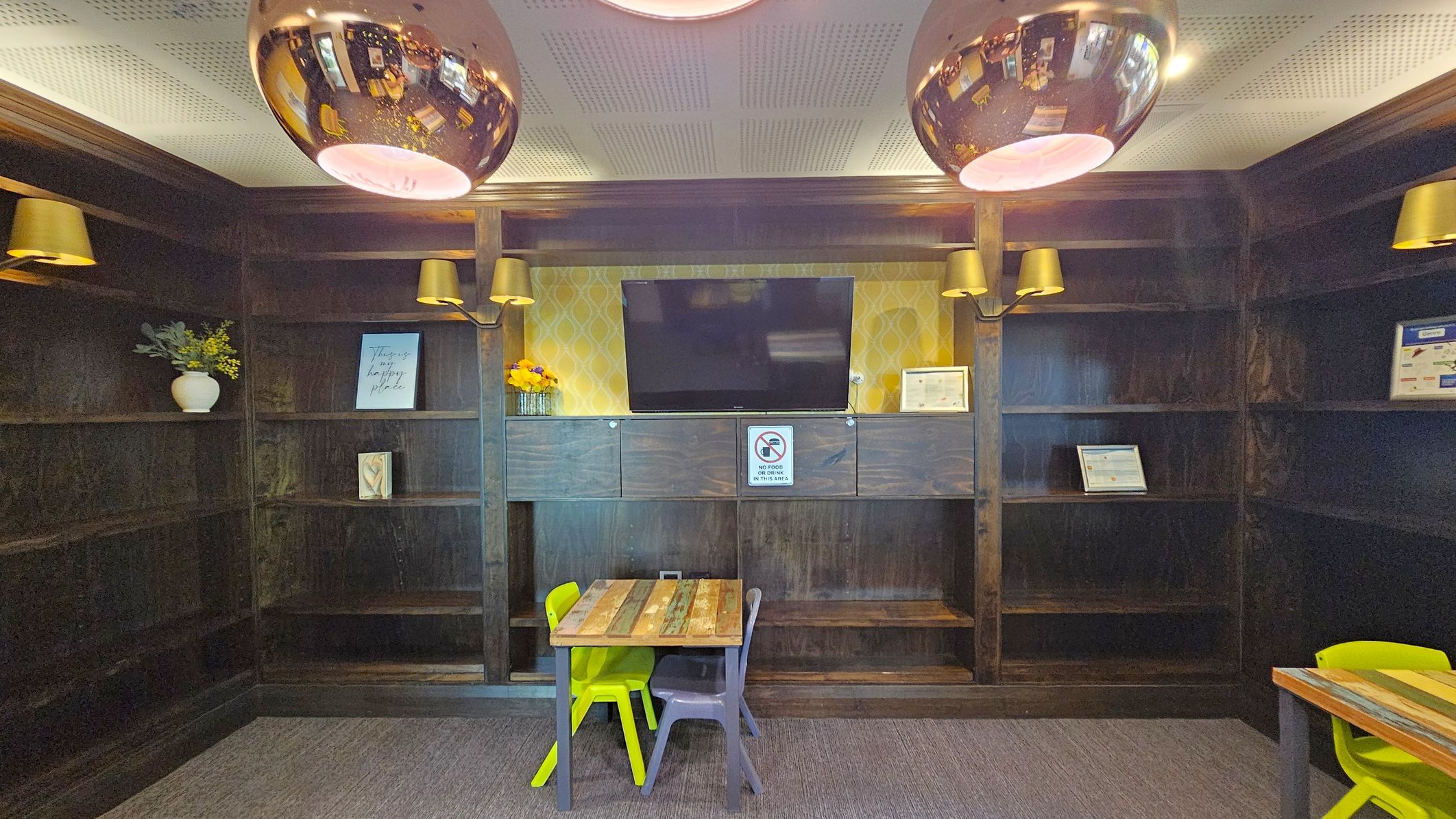 A room with dark wooden shelves lining the walls. In the centre, a flat-screen TV is mounted above a cabinet with a "No Food" sign. The shelves hold a few decorative items, including a vase with flowers and framed pictures. Below the TV, a small wooden table is accompanied by a yellow and a grey chair. The ceiling features perforated panels and two large, reflective pendant lights. The floor is carpeted in a neutral tone.