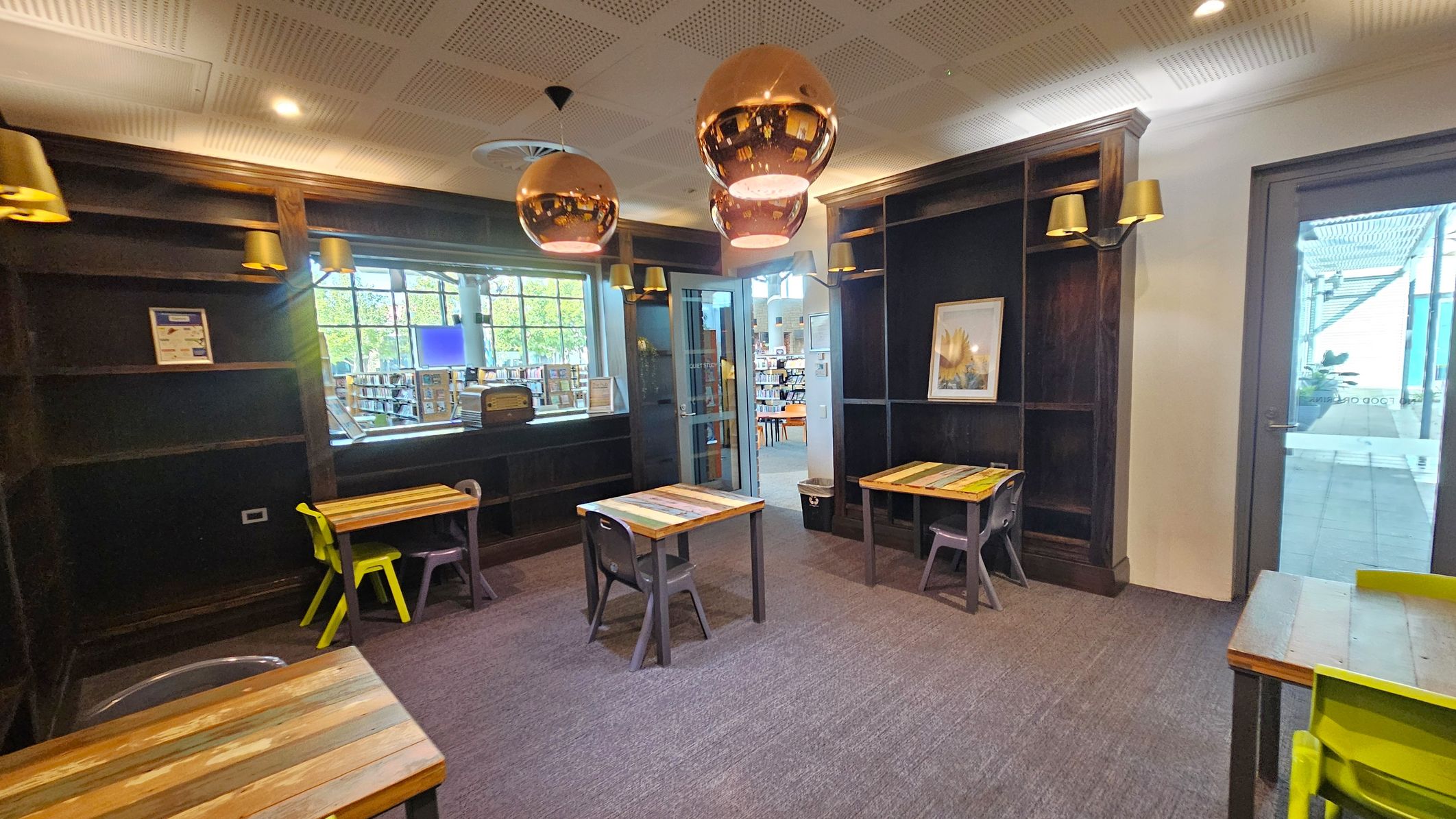 A room with dark wooden shelves on the left and right walls, featuring golden wall lamps. In the centre, there are three small tables with colourful wooden tops, each accompanied by a chair. The floor is carpeted in grey. Large copper pendant lights hang from the ceiling. A window and glass door on the back wall reveal a view of a library area with bookshelves. A framed picture is mounted on the right wall.