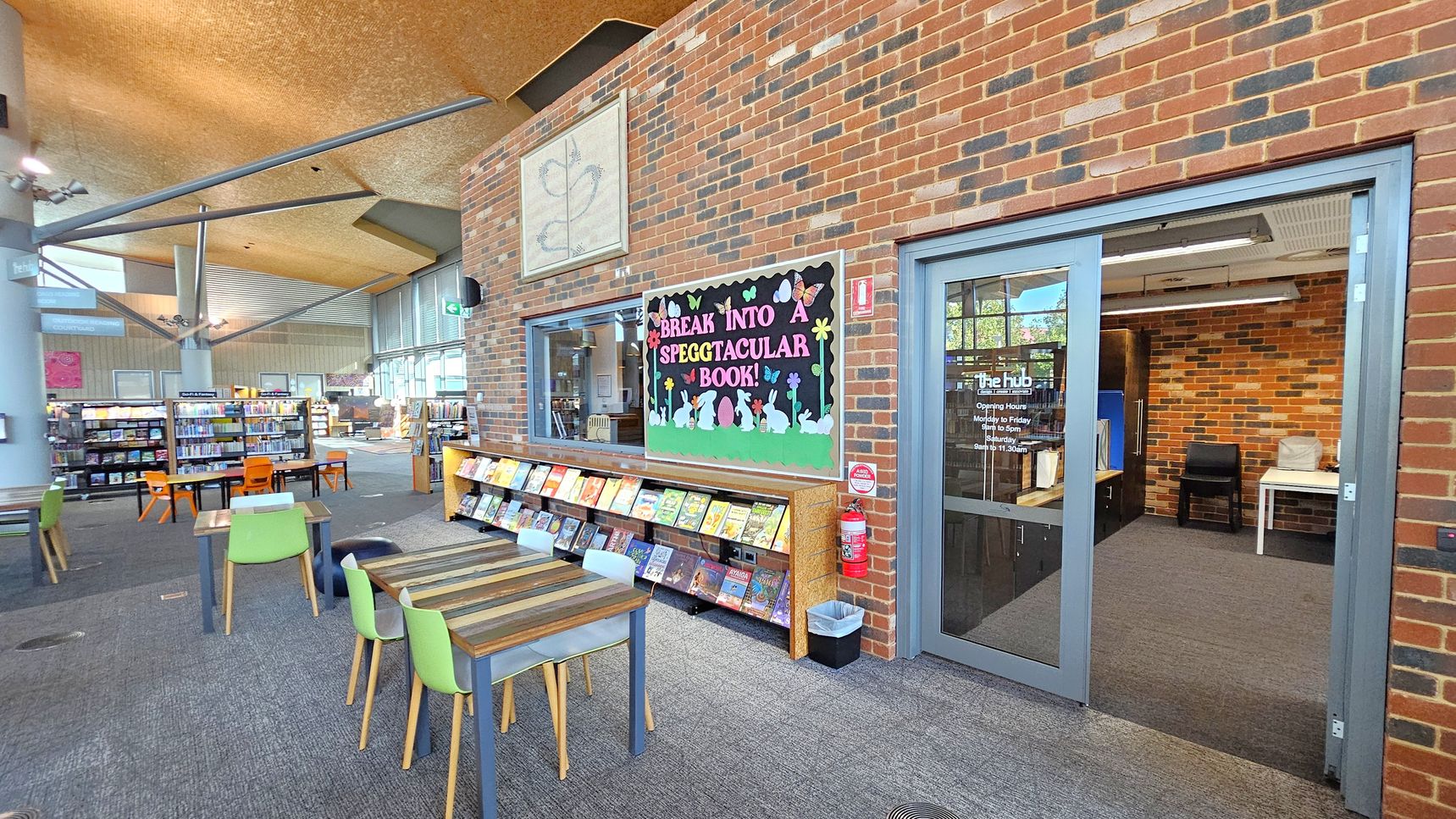 A library interior with a brick wall on the right featuring a colourful bulletin board that reads "Break into a Speggtacular Book!" Below it, a shelf displays various books. A glass door labelled "The Hub" leads to a room with a chair and table. On the left, there are tables with green and orange chairs, and bookshelves filled with books. The ceiling is high with exposed beams, and the floor is carpeted. A fire extinguisher and a waste bin are near the door.