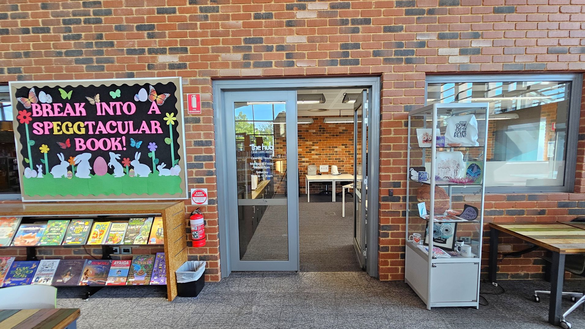 A brick wall with a colourful bulletin board on the left, featuring the phrase "Break into a Speggtacular Book!" with rabbits, eggs, and butterflies. Below, a shelf displays various books. A fire extinguisher and a waste bin are nearby. In the centre, a glass door leads to a room with tables and chairs. On the right, a glass display case contains various items, including mugs and decorative pieces. The floor is carpeted, and the ceiling has visible lighting fixtures.