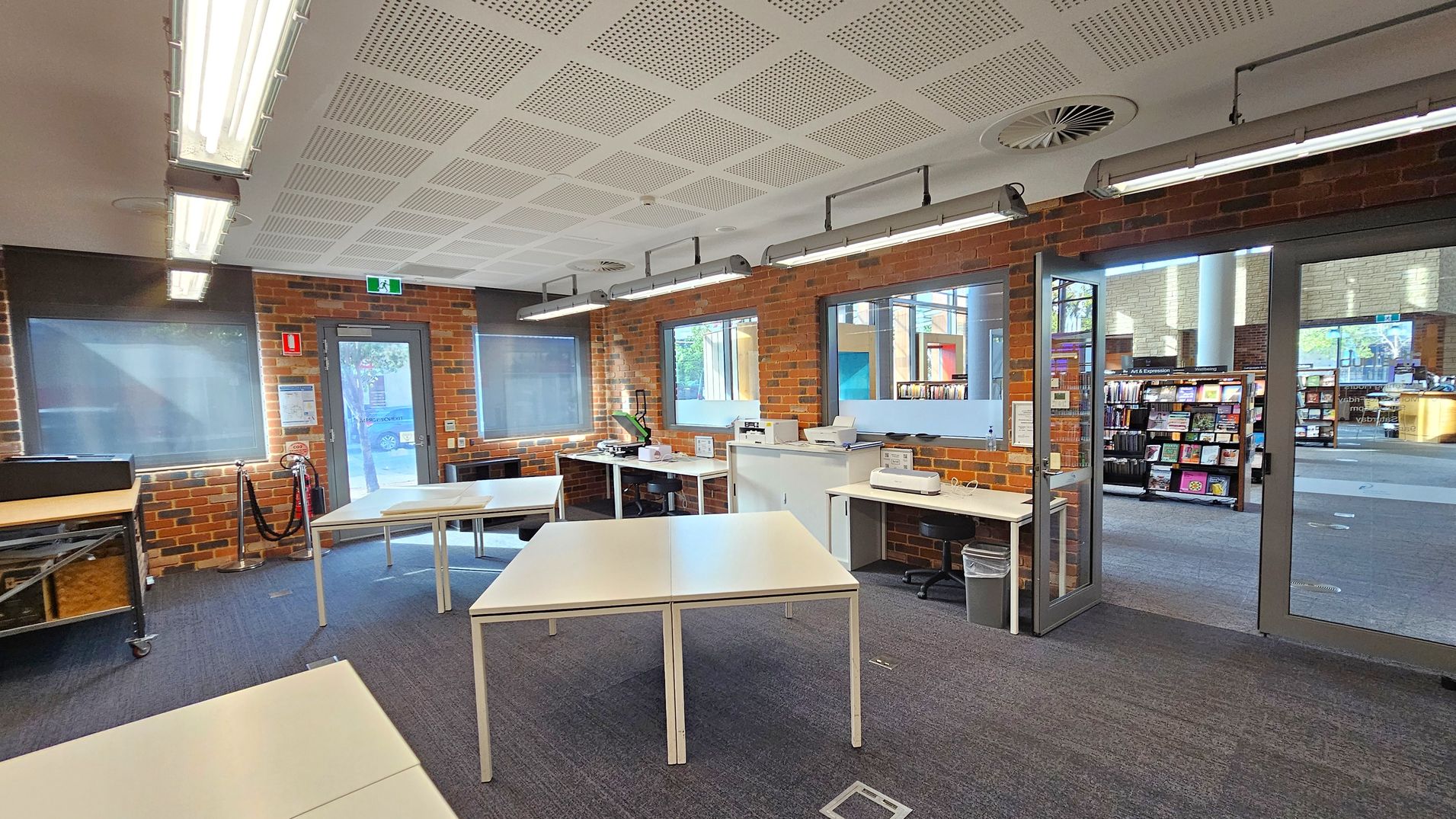 A room with brick walls and a grey carpeted floor. On the left, there are tables and a fire extinguisher. In the centre, white tables are arranged in a square formation. The right side features a glass door leading to a library area with bookshelves. The ceiling has white panels with fluorescent lights. Windows with blinds are on the left and centre walls, and there are various office machines on the tables.