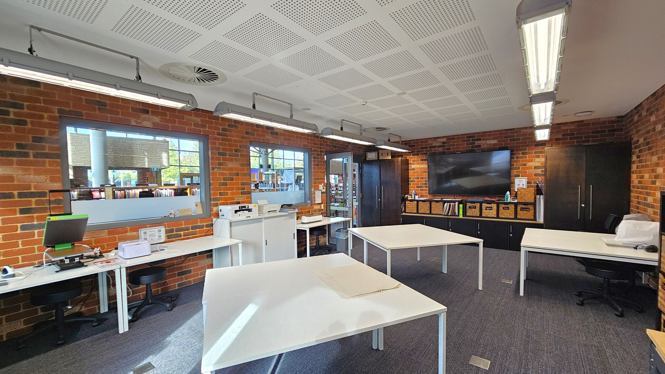 A room with brick walls and a perforated white ceiling. On the left, there are desks with computers and office equipment. In the centre, several white tables are arranged with a clear workspace. The right side features a large flat-screen TV mounted on the wall above a row of cabinets with labelled storage boxes. Fluorescent lights are suspended from the ceiling, and windows provide a view into another room with bookshelves. The floor is carpeted in grey.