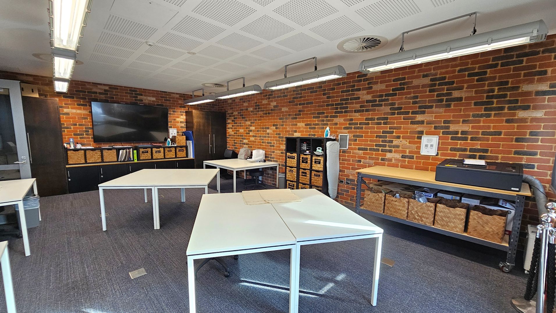 A room with brick walls and a grey carpeted floor. On the left, a large flat-screen TV is mounted above a cabinet with wicker baskets. In the centre, several white tables are arranged in a square formation. On the right, a shelving unit holds more wicker baskets and an ironing board. Above, fluorescent lights are suspended from a white perforated ceiling.