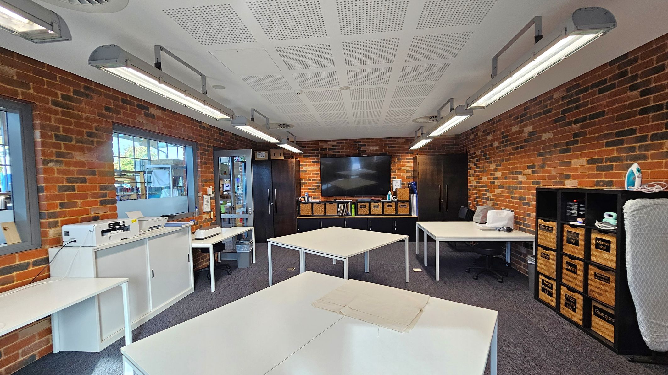 A room with brick walls and a grey carpeted floor. On the left, there are white cabinets with printers and a window. In the centre, several white tables are arranged, and a large flat-screen TV is mounted on the wall above a cabinet with wicker baskets. On the right, a shelving unit holds labelled baskets and an iron. The ceiling is white with recessed lighting and ventilation.