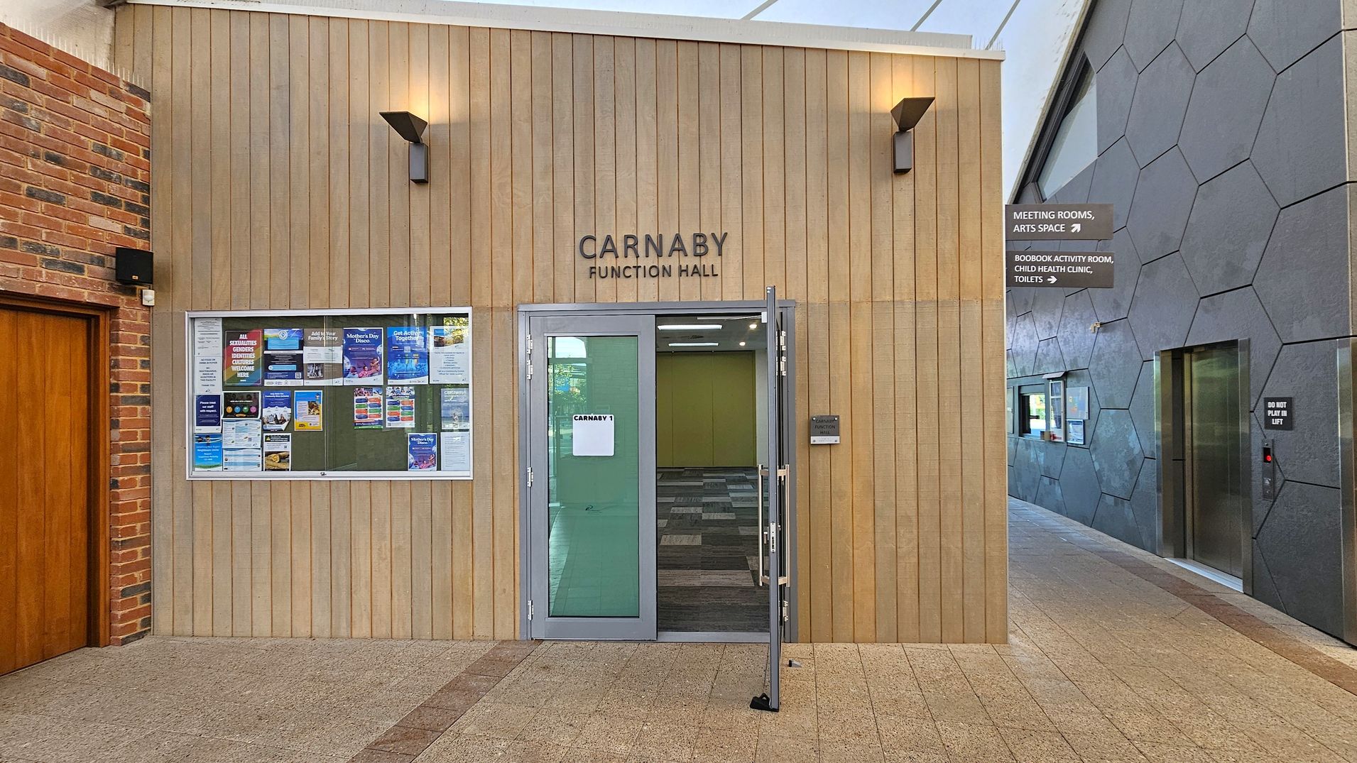 Carnaby Function Hall entrance with a light wood-panelled facade. A glass door is slightly open, revealing a carpeted interior. To the left, a notice board displays various posters. The floor is tiled in a light brown shade. On the right, a grey wall with hexagonal patterns features an elevator and directional signs for meeting rooms and other facilities. The ceiling is white and slightly visible.