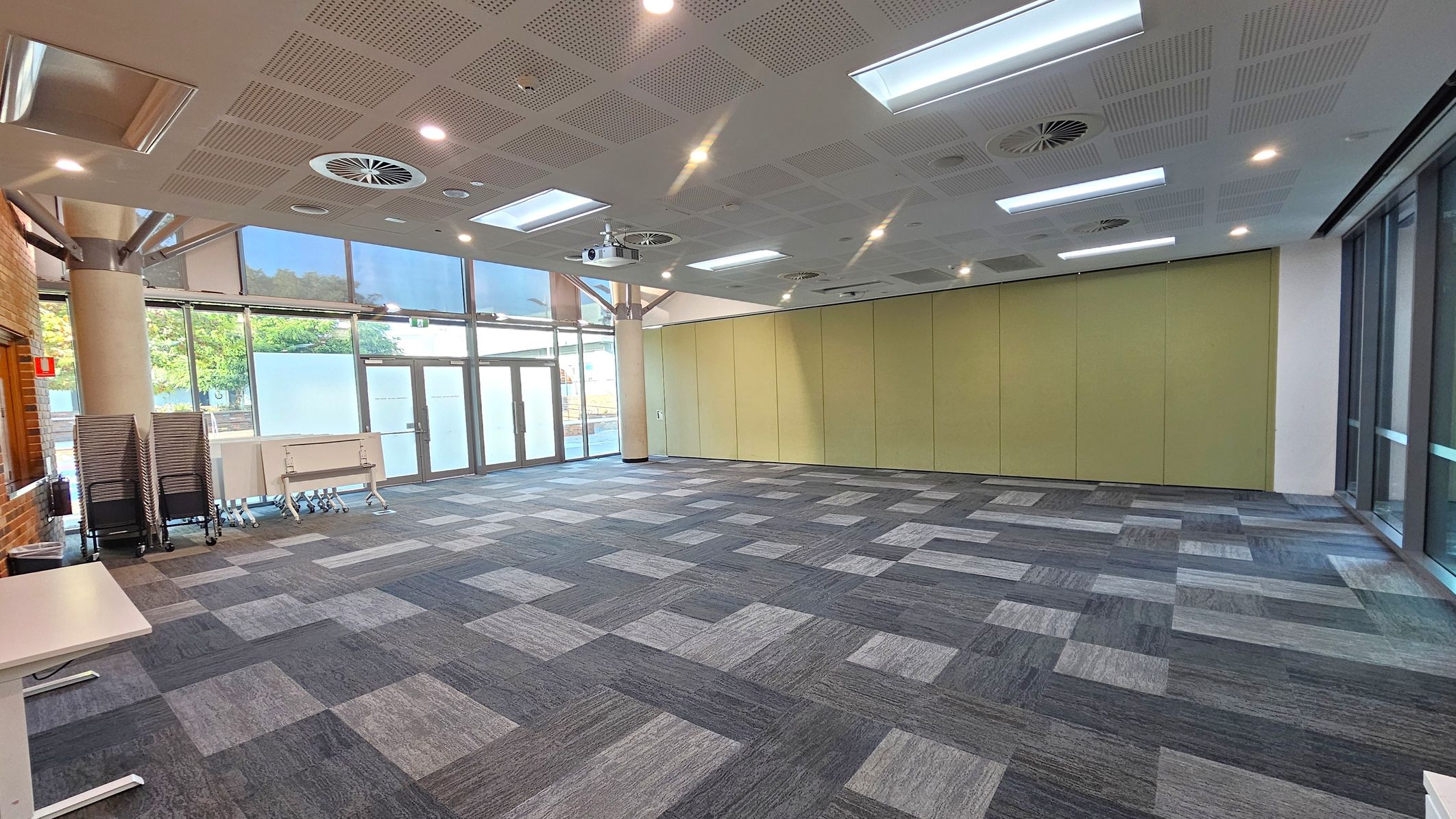 A spacious room with a patterned carpet of grey and blue squares. On the left, there are stacked chairs and folded tables against a brick wall. The right side features large windows with frosted glass panels. The back wall has light green partition panels. The ceiling is fitted with recessed lights, air vents, and a projector.