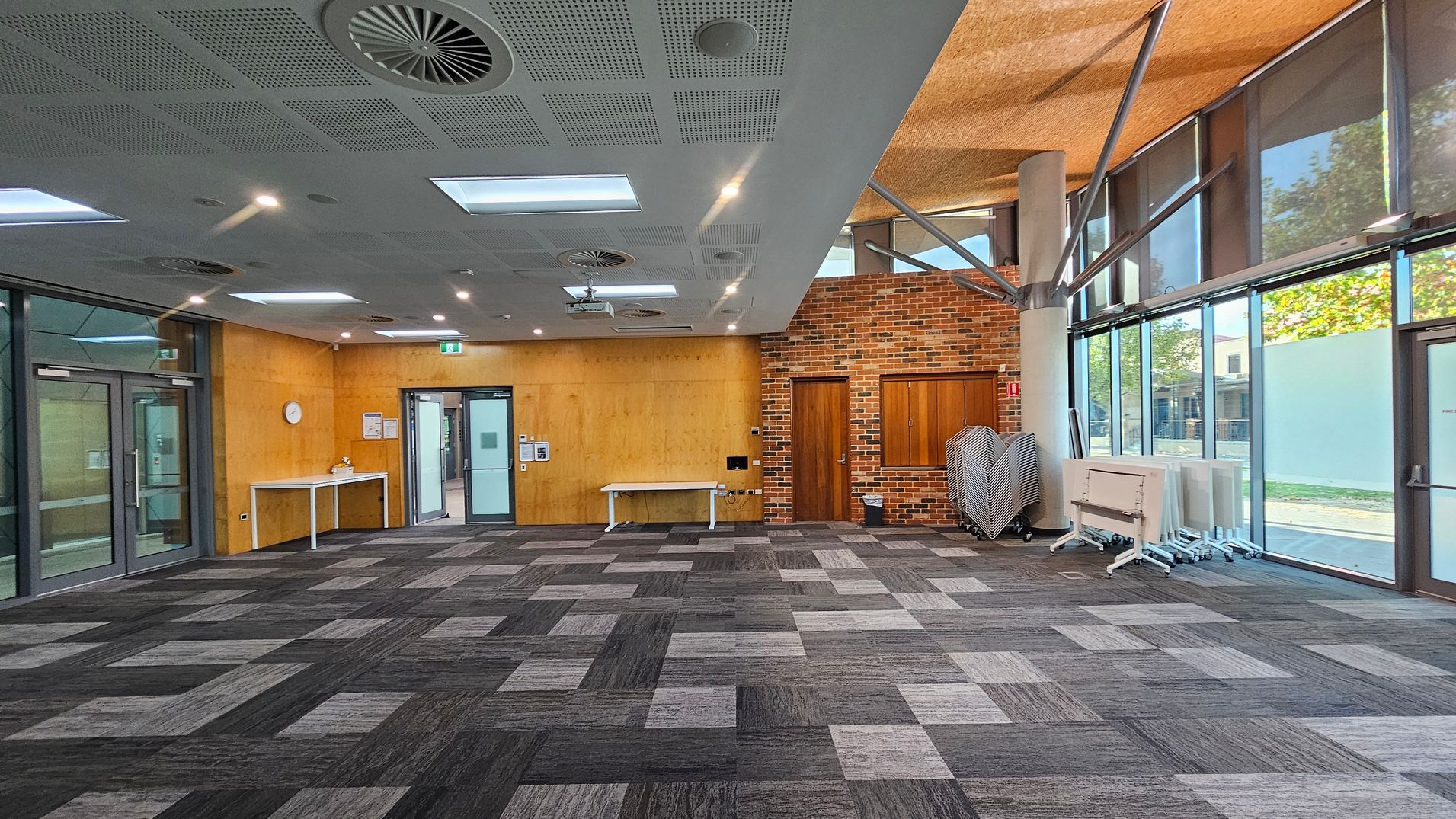 A spacious room with a patterned carpet floor in shades of grey and purple. The left side features a wooden wall with a clock and a white table. The centre has a doorway and another white table against a wooden wall. The right side has a brick wall with a wooden door and folded tables and chairs stacked near large windows. The ceiling is fitted with lights and air vents.