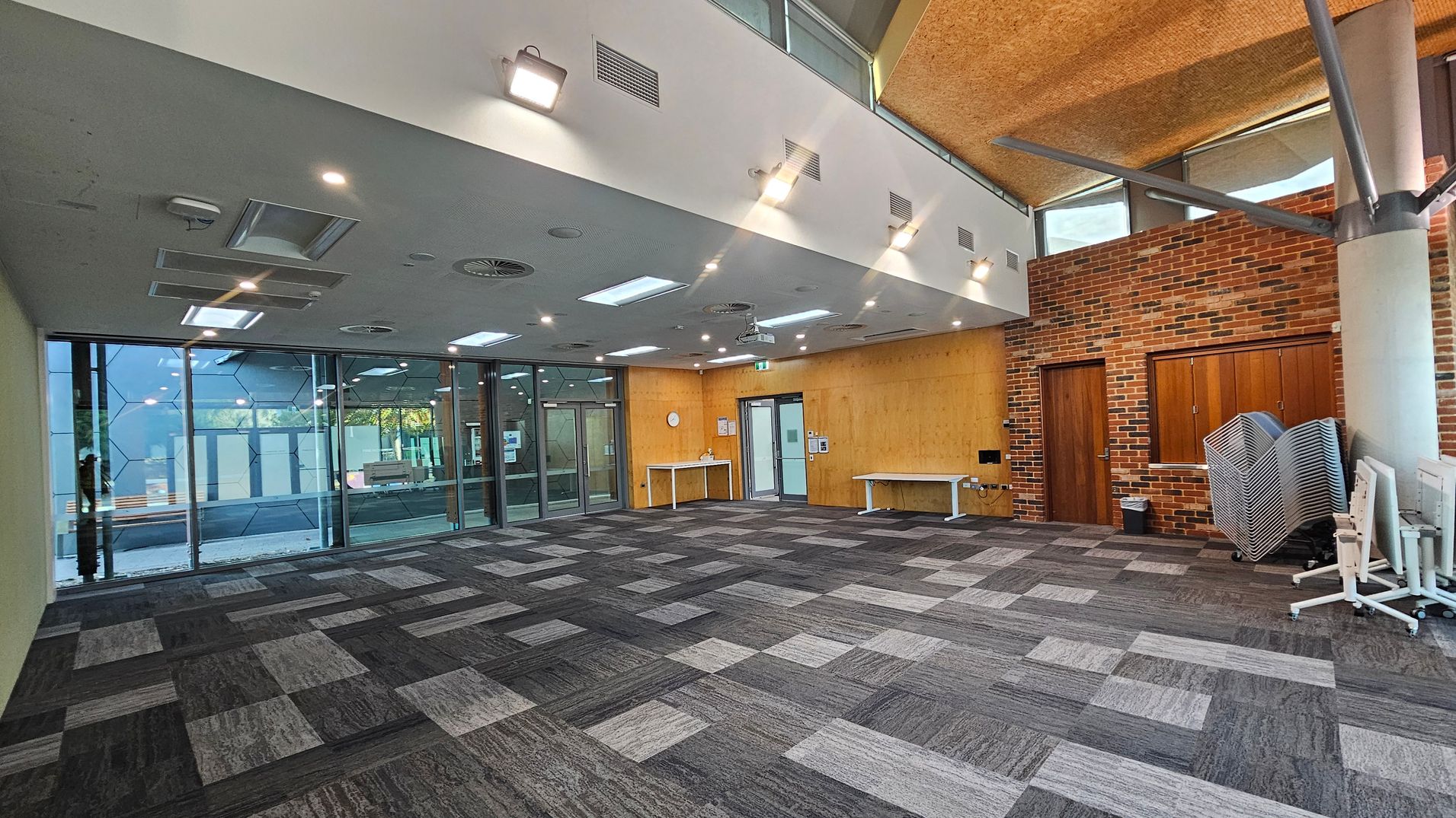 A spacious room with a patterned carpet floor in shades of grey. The left side features large glass windows with a geometric design, allowing natural light to enter. The right side has a brick wall with two wooden doors and a stack of folded chairs. The ceiling is white with recessed lighting and ventilation panels. A wooden wall with a clock and two white tables is at the back. The ceiling has a section with a textured finish and exposed beams.