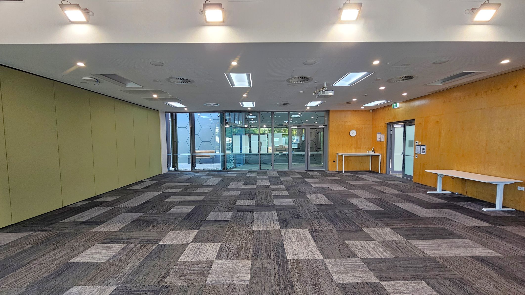 A spacious room with a patterned carpet in shades of grey and pink. The left wall is green, while the right wall is wooden with a clock and a white table. The back wall features large glass doors and windows with a geometric pattern. The ceiling is white with recessed lighting and air vents. Two white tables are positioned against the right wall.