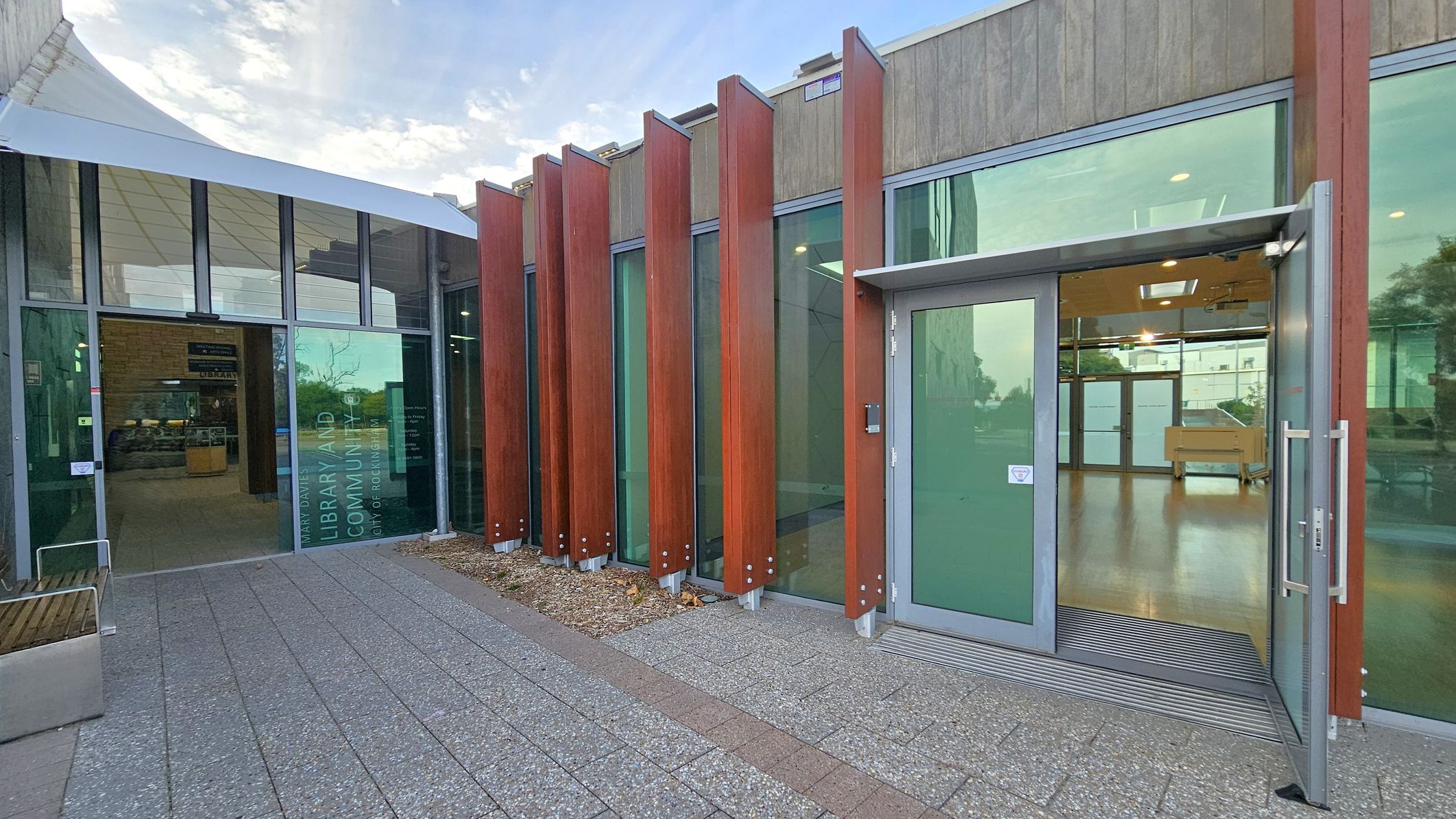 A modern building entrance with large glass panels and vertical wooden slats on the right. The door is open, revealing a spacious interior with wooden flooring. On the left, a glass facade with text indicating a library and community centre. The floor outside is paved with textured tiles, and a white canopy extends over part of the entrance.