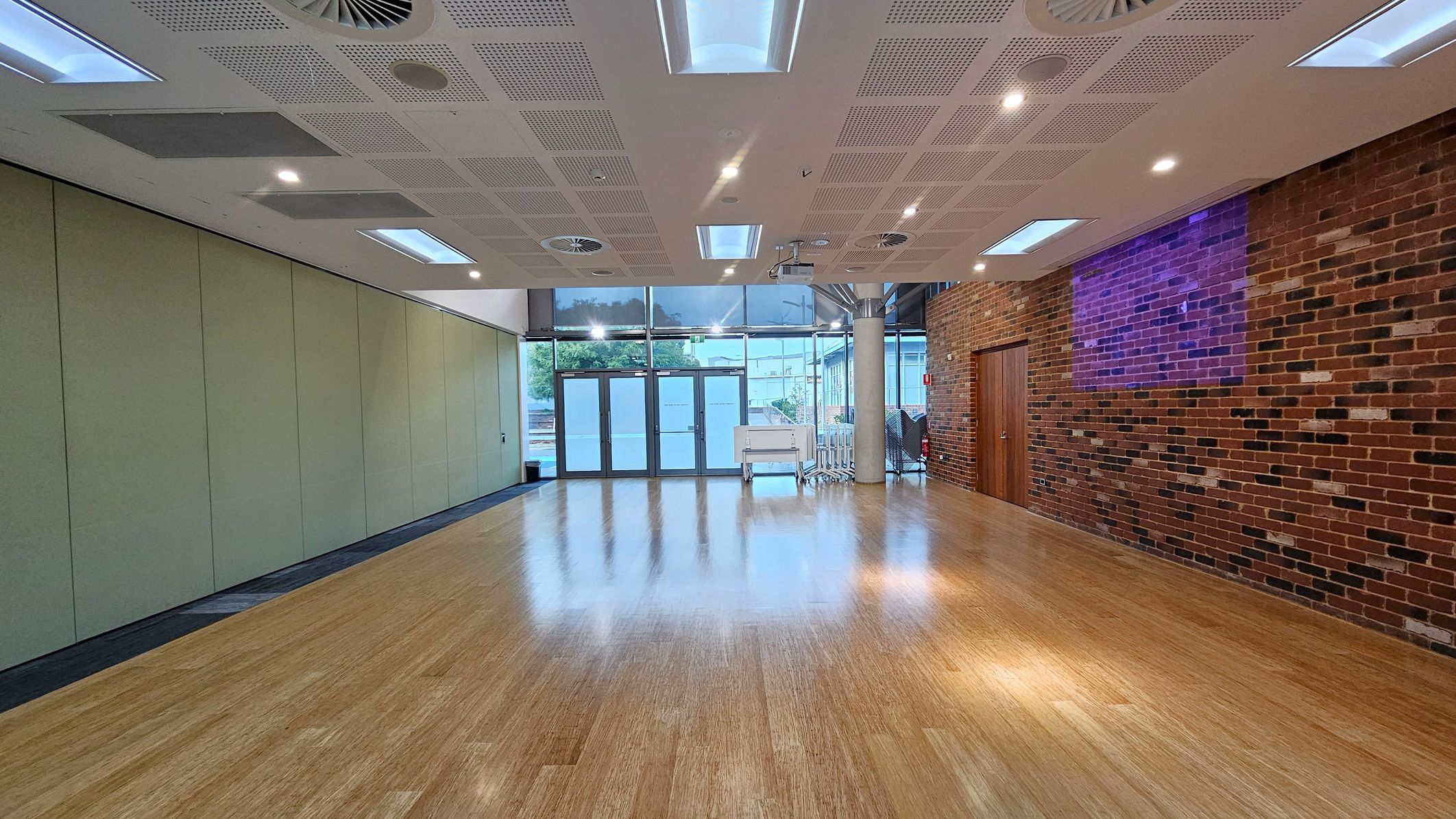 A spacious room with a wooden floor and a ceiling featuring recessed lighting and ventilation panels. On the left, there is a wall with light green panels. The right side has a brick wall with a wooden door. At the far end, large glass doors and windows allow natural light in, with a view of trees outside. A column and some stacked chairs are near the glass doors.