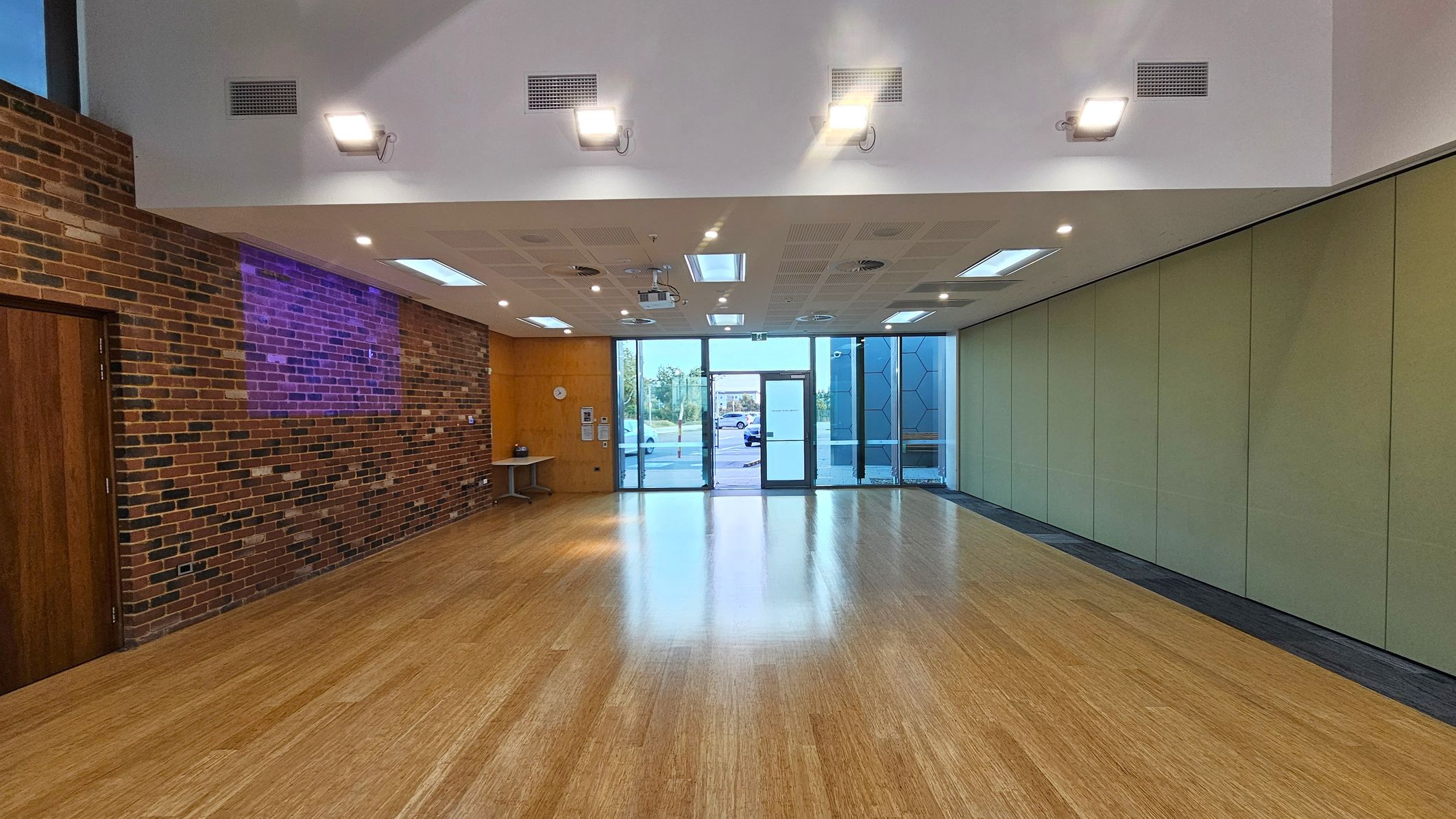 A spacious room with a wooden floor and a high ceiling featuring recessed lighting. On the left, there's a brick wall with a wooden door and a small table. The right side has a series of greenish partition panels. The back wall is mostly glass, with large windows and a central glass door leading outside. The ceiling has multiple light fixtures and ventilation grilles.