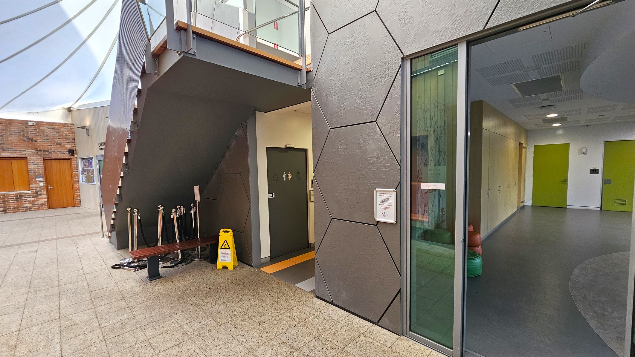 A modern indoor space with a geometric wall design on the right, featuring hexagonal patterns. A glass door reveals a hallway with green doors and a grey floor. On the left, a staircase with a metal railing leads upwards. Below the stairs, a door marked with restroom symbols is visible. A yellow caution sign is placed near the entrance. The floor is tiled, and a brick wall with wooden doors is in the background. A glass ceiling allows natural light to illuminate the area.