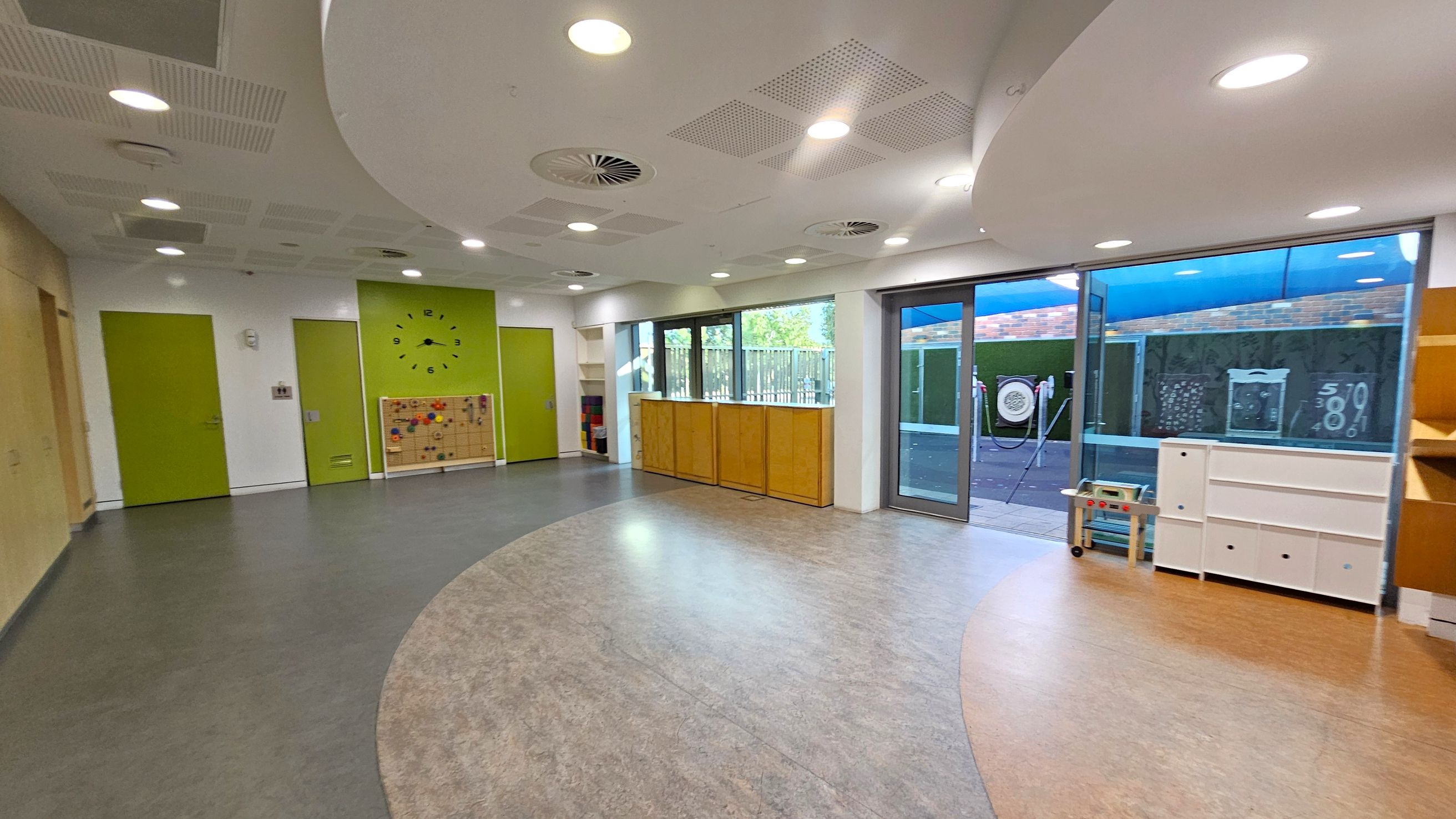 A spacious room with a curved, two-toned floor in grey and beige. On the left, there are green doors and a wall with a large clock and a pegboard with colourful pegs. The right side features large windows and glass doors leading outside, with wooden cabinets below. A small play kitchen and white storage units are near the windows. The ceiling is white with circular lights and ventilation units.