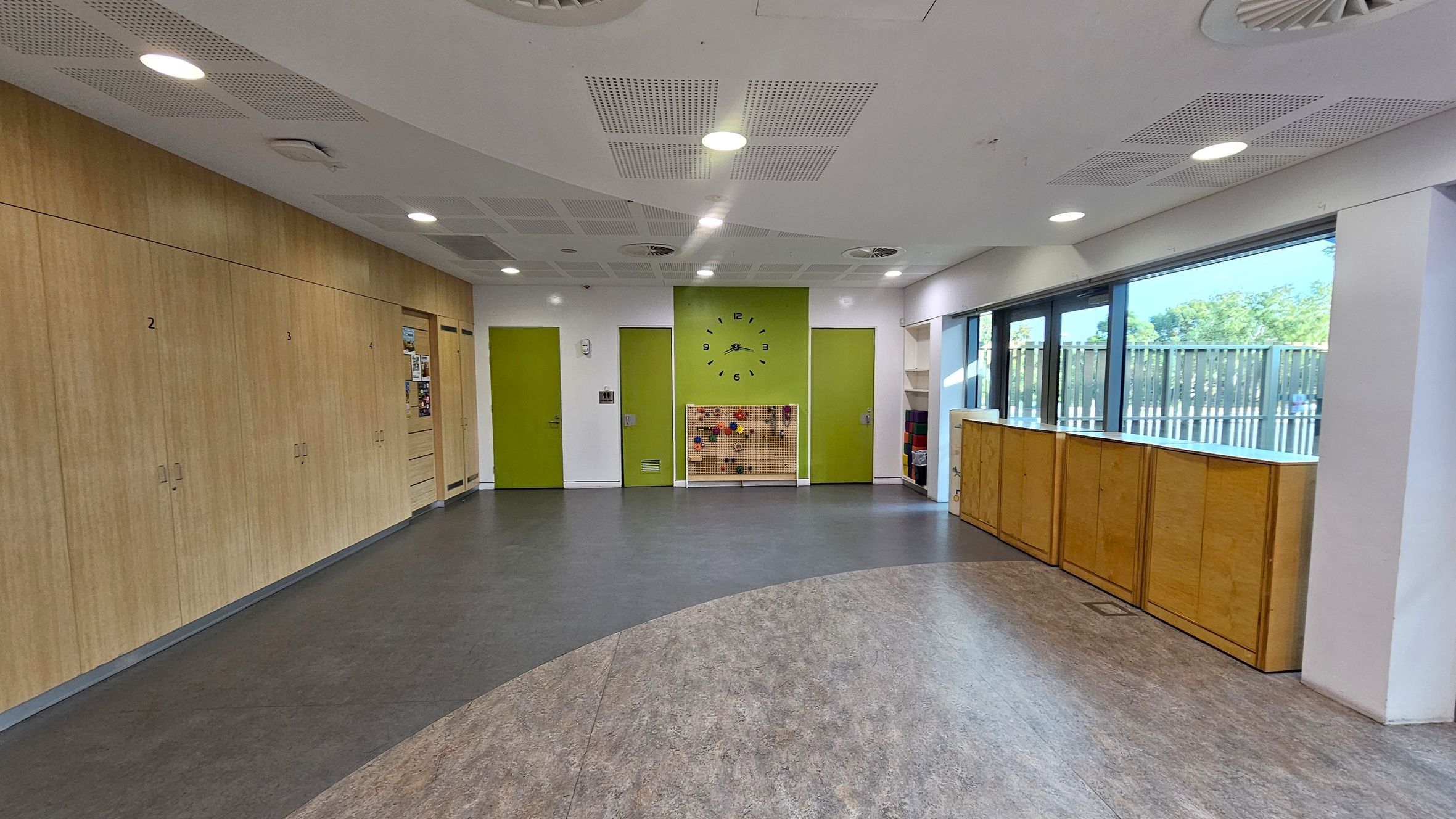 A spacious room with a grey and brown patterned floor. On the left, there are wooden cabinets numbered 2 and 3. The back wall features two green doors and a white door, with a large clock above a pegboard with colourful items. On the right, large windows allow natural light in, with wooden cabinets beneath them. The ceiling is white with recessed lighting and ventilation panels.