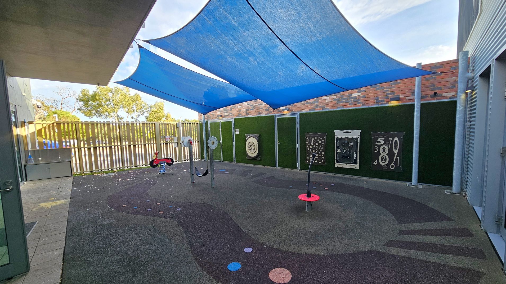 A children's play area with a soft, dark rubber floor featuring colourful circular patterns. In the centre, there are play structures, including a red spring rider and a spinning element. The area is shaded by large blue fabric canopies. The background has a green wall with interactive panels and a brick wall above. On the left, there's a wooden fence with trees visible beyond it. The ceiling is formed by the blue canopies, and the floor is a mix of dark and light grey rubber.