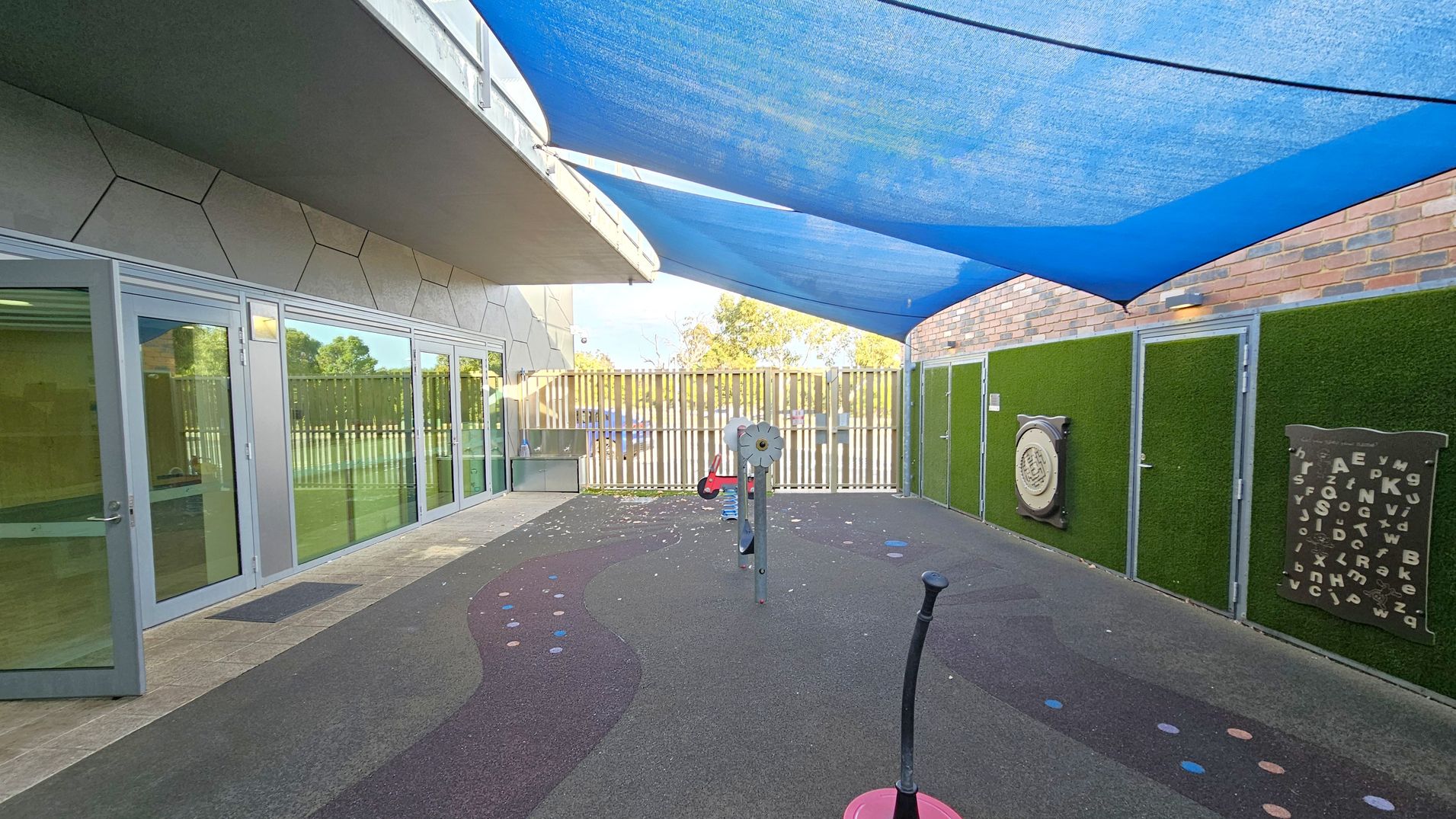 Outdoor play area with a rubberised, multicoloured floor featuring circular patterns. On the left, a building with large glass doors and hexagonal wall panels. The right side has a green artificial grass wall with educational boards displaying letters and numbers. A blue shade sail covers the area. In the centre, play equipment is visible. The background shows a wooden fence with trees beyond.