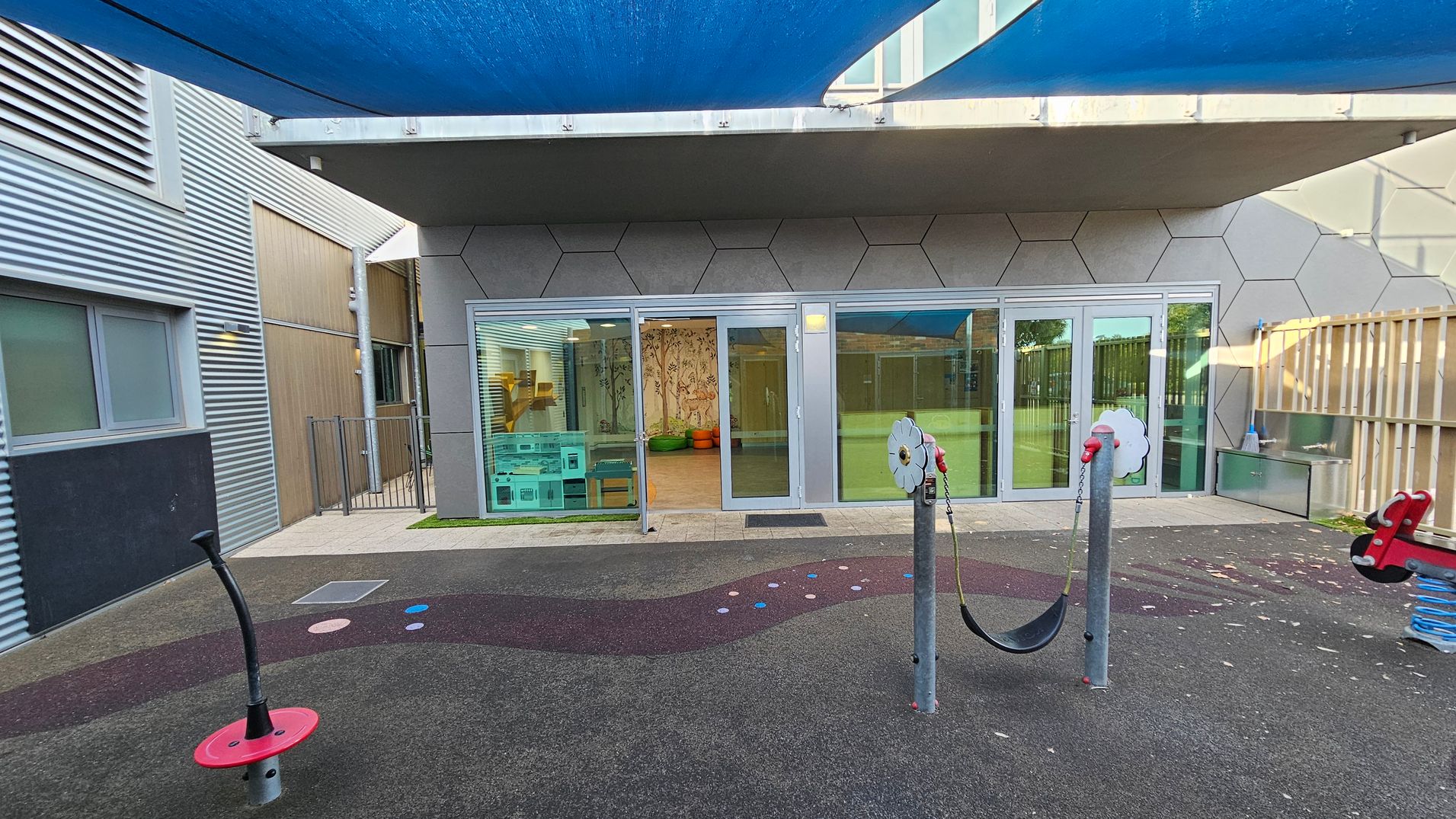 A playground area with a rubberised surface featuring a wavy pattern and colourful dots. In the foreground, there are play structures including a red spinning pole and a swing. The background shows a building with large glass doors and hexagonal wall patterns. Inside, there are colourful cushions and a mural. The left side has a corrugated metal wall with windows, while the right side has a wooden fence. A blue shade cloth is overhead.