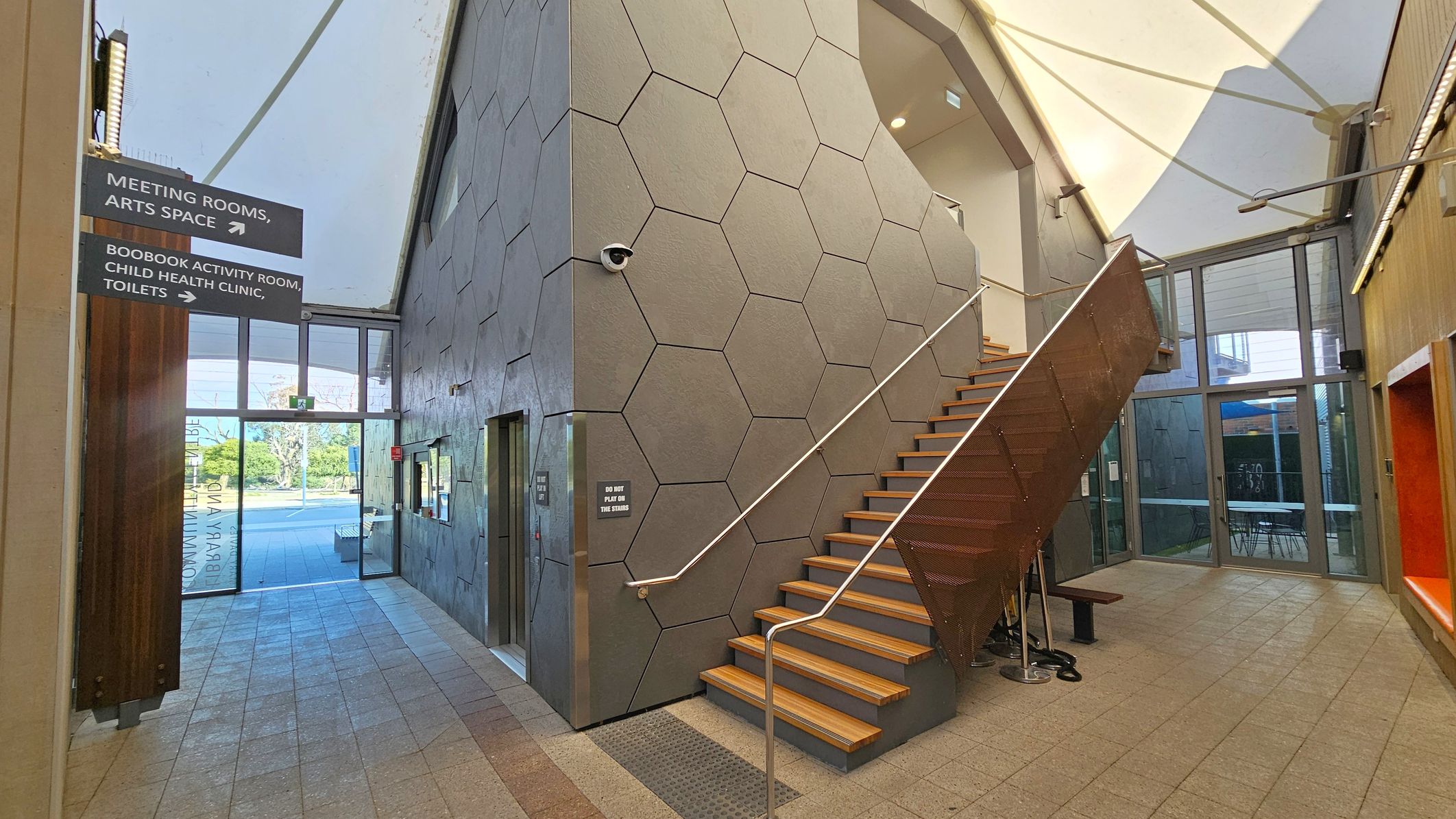 A modern interior with a hexagonal-patterned wall in the centre, featuring a staircase with wooden steps and a metal railing. To the left, signs indicate directions to meeting rooms, an arts space, and other facilities. The floor is tiled, and there is a glass entrance with a view of greenery outside. On the right, a seating area with an orange bench is visible. The ceiling has a white, tent-like structure with lights.