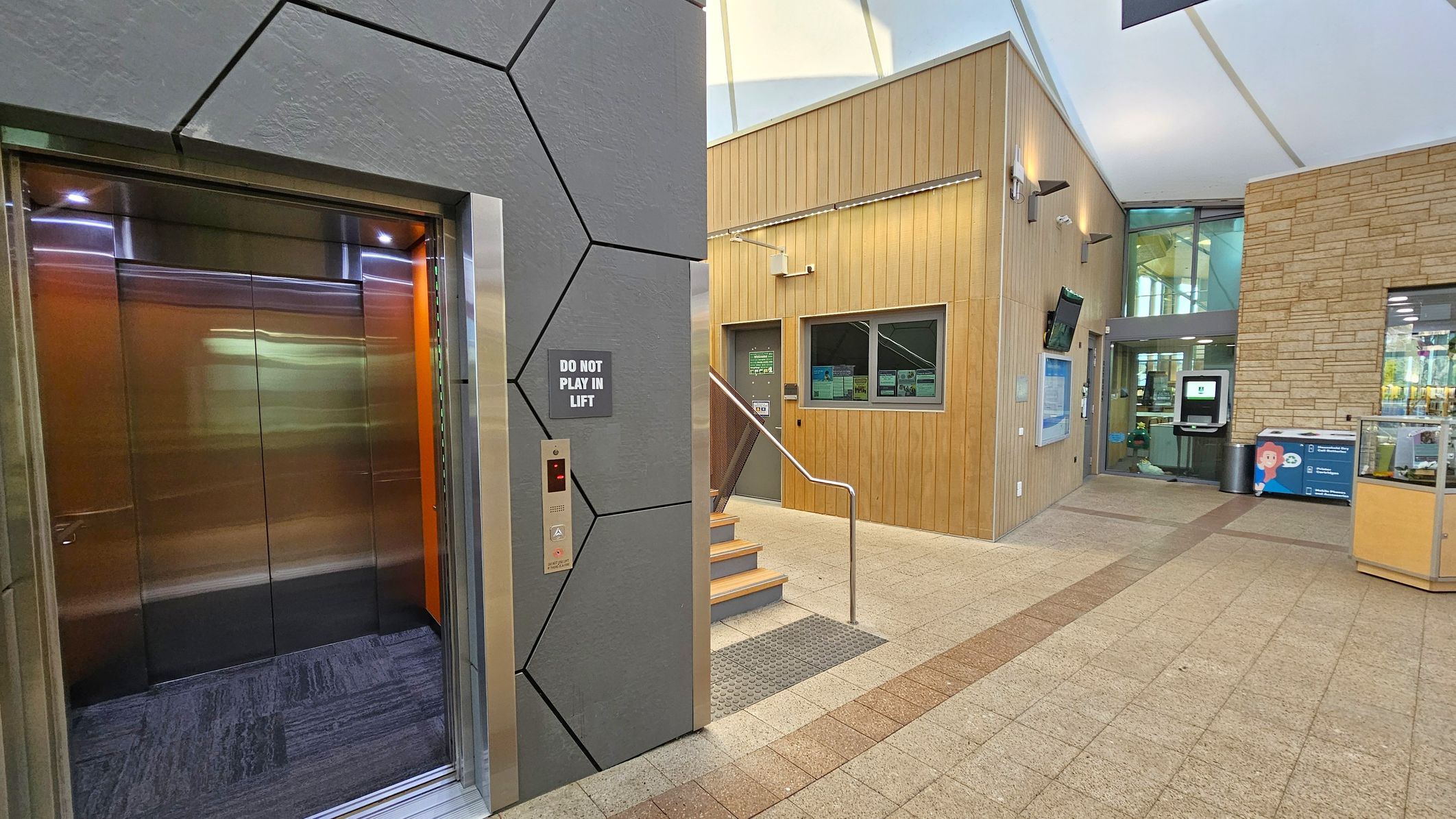 On the left, there is an open lift with metallic doors and a sign that reads "DO NOT PLAY IN LIFT". The wall around the lift has a geometric pattern. In the centre, a small set of stairs with a metal railing leads up to a wooden-panelled wall with a door and window. On the right, there is a glass entrance with a digital kiosk and a stone wall. The floor is tiled with a mix of beige and brown tiles, and the ceiling is white with a modern design.