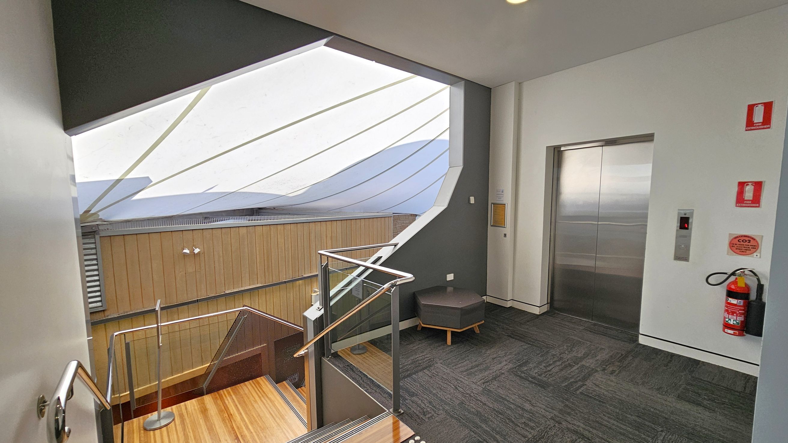 A modern indoor space with a staircase on the left featuring wooden steps and a metal railing. The right side has a lift with a metallic door, surrounded by white walls. A fire extinguisher is mounted on the wall next to the lift. The floor is carpeted in a dark grey pattern. Above, a large, angled skylight with white panels allows natural light to illuminate the area. A small, hexagonal stool is placed near the lift.