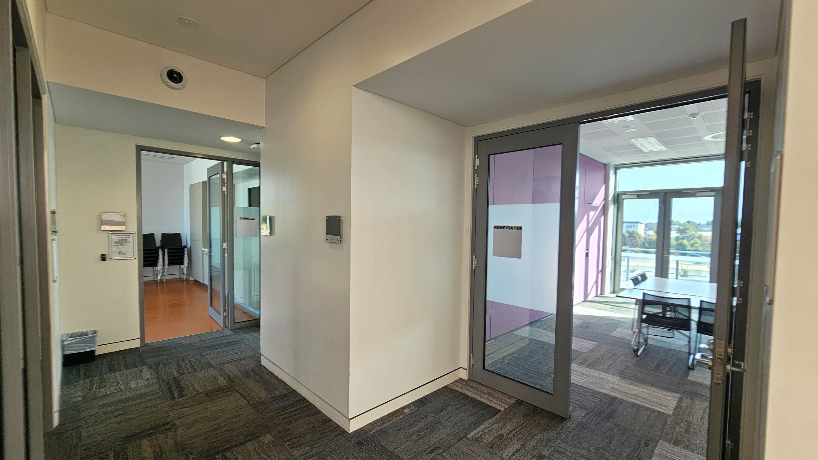 A hallway with a patterned carpet floor leads to two rooms. On the left, a room with a wooden floor and stacked chairs is visible through a glass door. On the right, another room with a glass door has a table and chairs, with large windows allowing natural light in. The walls are light-coloured, and the ceiling has recessed lighting and a security camera.