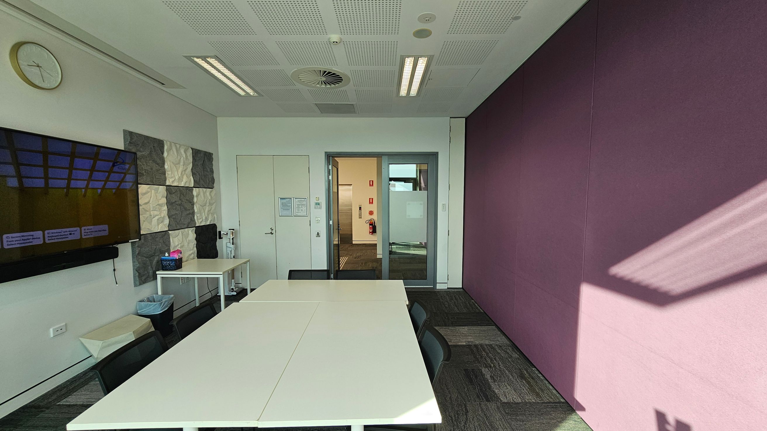 A meeting room with a white table and black chairs in the centre. On the left wall, there's a clock and a mounted TV above a small table with a basket. The wall has textured panels in grey and white. A door and a glass panel lead to another room. The right wall is covered in a purple fabric, with sunlight casting shadows. The ceiling is white with recessed lights and ventilation. The floor is carpeted in a grey pattern.