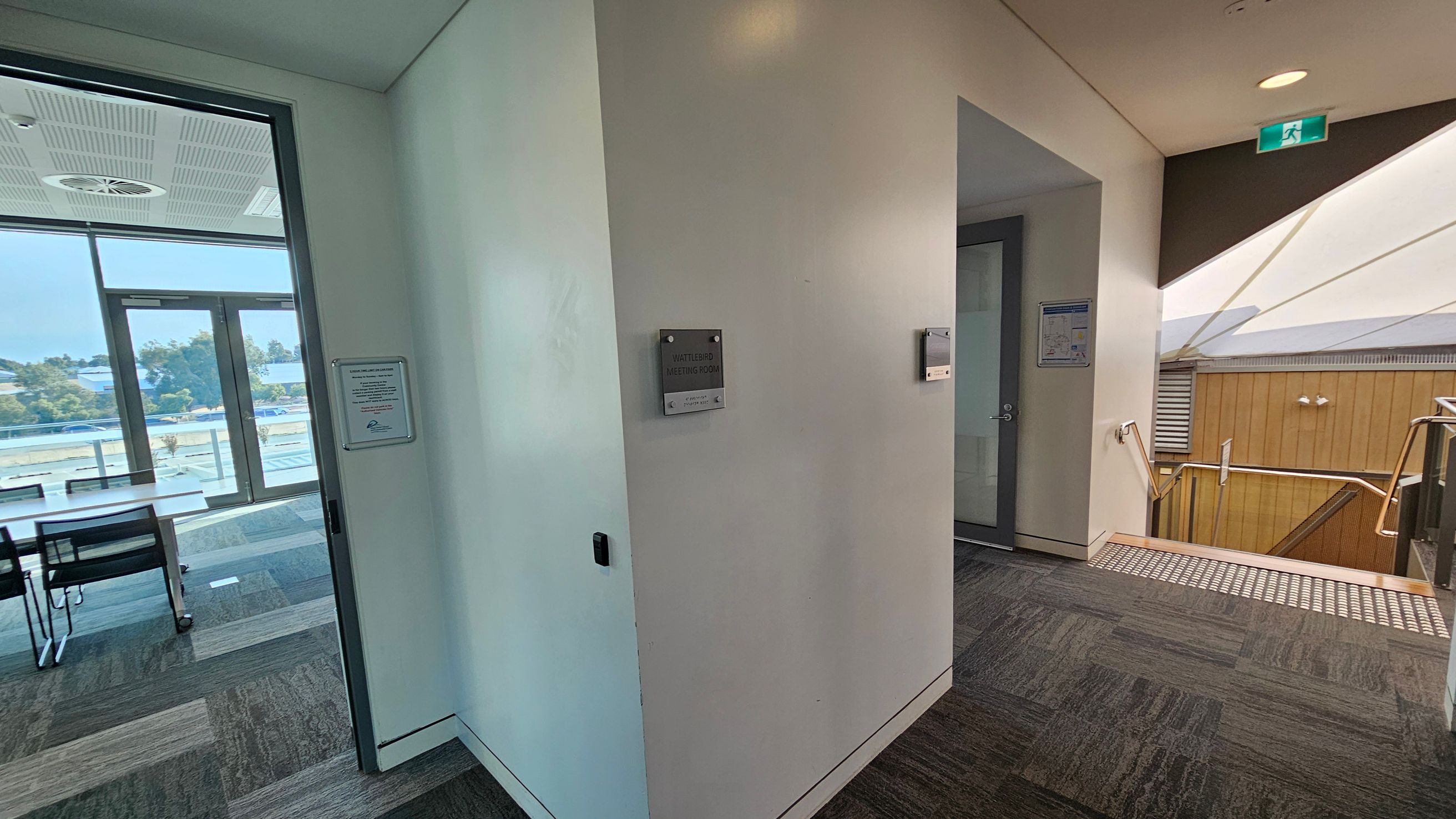 A corridor with a meeting room on the left, visible through a glass door with a sign reading "Wattlebird Meeting Room". Inside, there are tables and chairs. The floor is carpeted with a multicoloured pattern. On the right, a stairway with metal railings leads downwards. The ceiling has recessed lighting and an emergency exit sign. The background shows large windows with a view of trees and buildings outside.