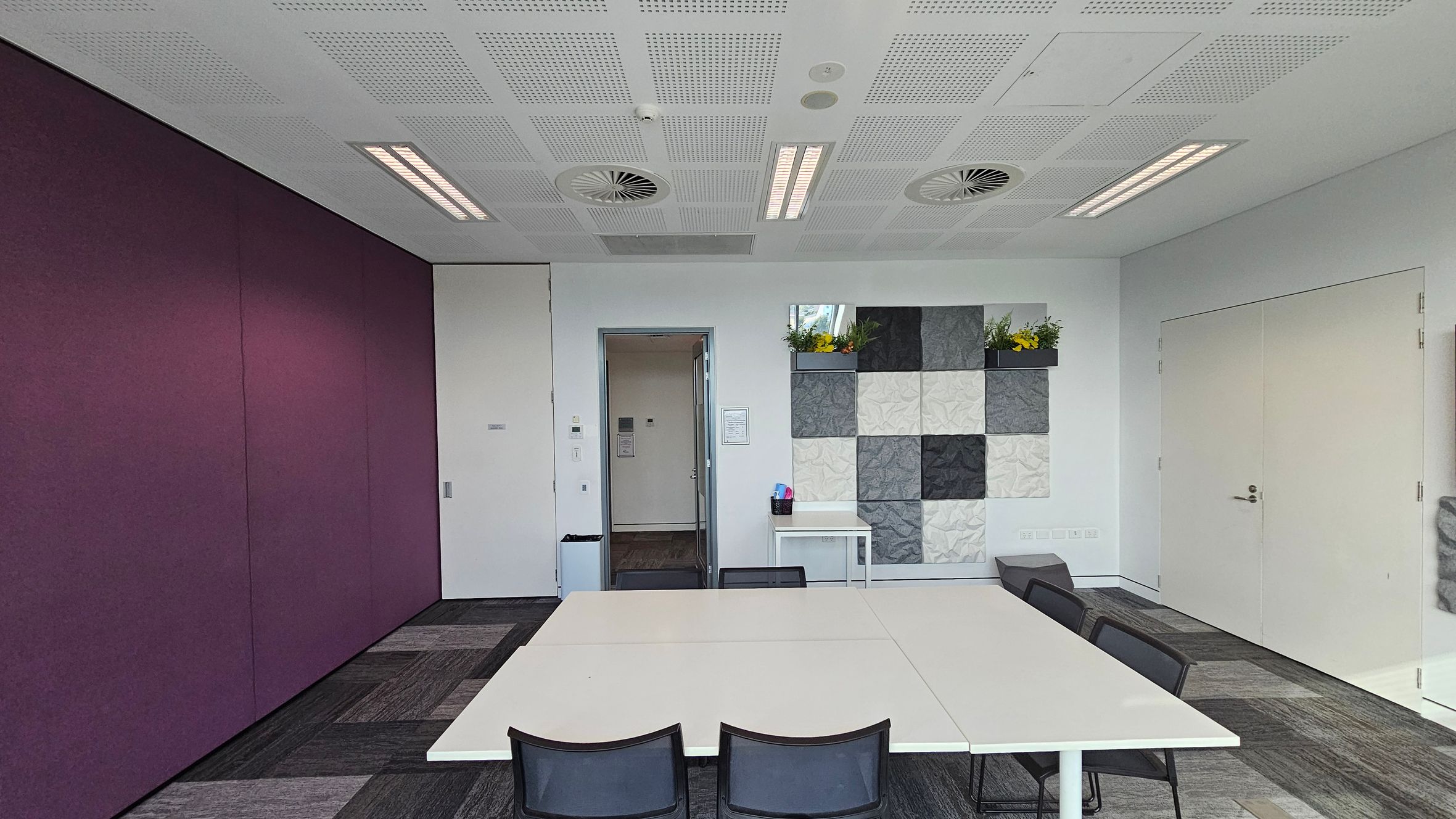 A modern room with a purple wall on the left and a white wall on the right. In the centre, there's a doorway leading to another room. A decorative wall with grey and white square panels is on the right, with a small shelf holding yellow flowers. Below it, a small table with office supplies. In the foreground, white tables and black chairs are arranged. The ceiling has recessed lighting and circular vents, and the floor is covered with grey carpet tiles.