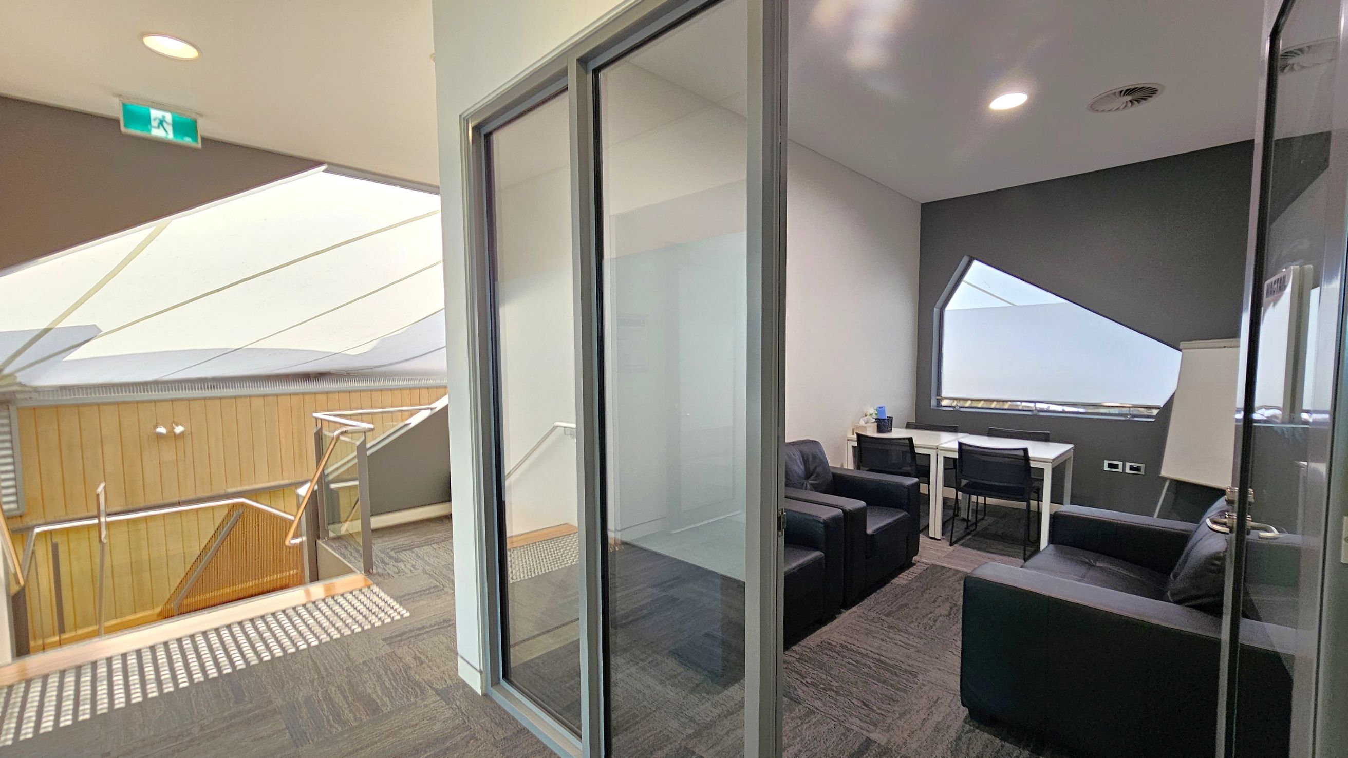 On the left, there is a staircase with a wooden railing and a textured metal floor panel. Above, a ceiling with recessed lighting and an emergency exit sign. On the right, a small meeting room with glass walls, containing two black armchairs, a white table with two chairs, and a flip chart. The room has a geometric window and a grey accent wall. The floor is carpeted with a patterned design.