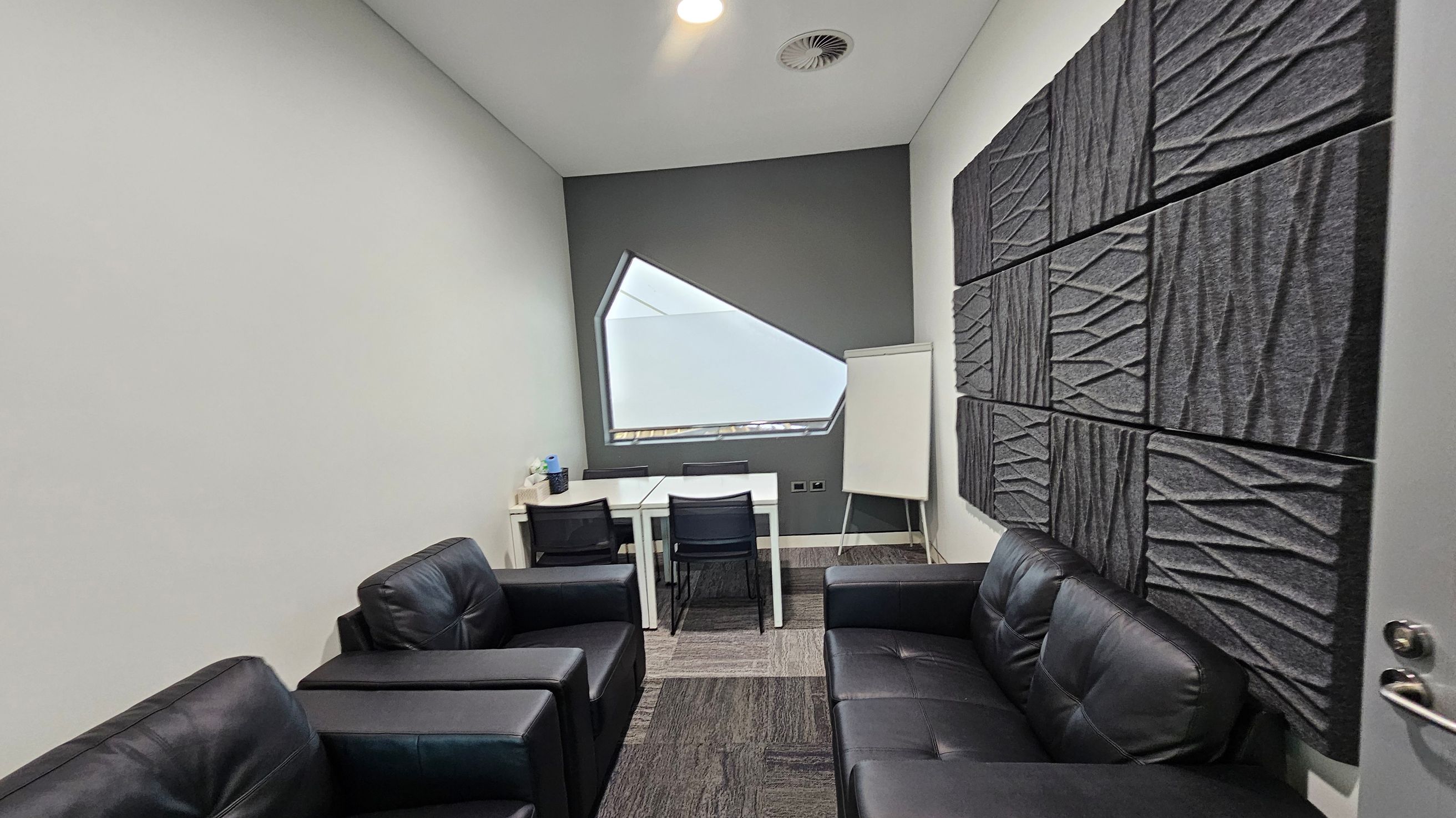 A small room with a modern design. On the left, there are two black leather armchairs. On the right, a black leather sofa is positioned against a wall with textured grey acoustic panels. In the middle, a white table with two black chairs faces a uniquely shaped window. A flip chart stands beside the table. The floor is carpeted with a mix of grey and patterned tiles, and the ceiling is white with a recessed light and air vent.