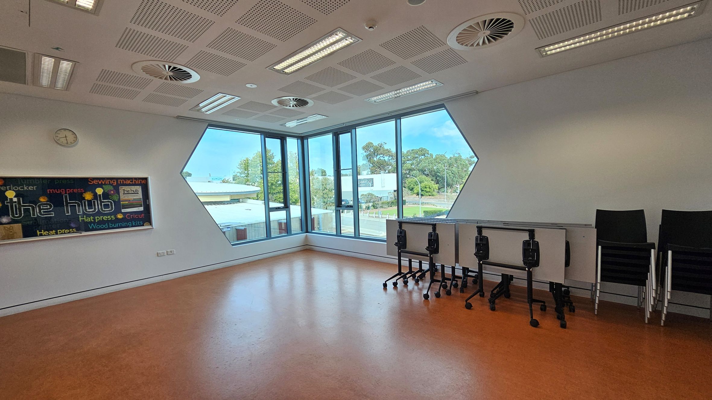 A room with orange flooring and a white ceiling featuring circular vents and rectangular lights. On the left wall, a colourful bulletin board titled "the hub" is displayed, with a clock above it. The right side has stacked black chairs and foldable tables on wheels. Large geometric windows on the far wall offer a view of trees and buildings outside.