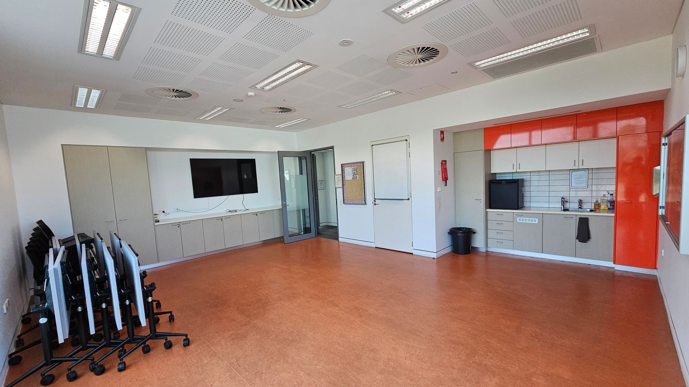 A room with an orange-brown floor and a white ceiling featuring multiple air vents and lights. On the left, there are stacked tables with wheels. The centre has a wall-mounted TV above a row of cabinets. On the right, there's a kitchenette with red and beige cabinets, a sink, and a small fridge. A fire extinguisher is mounted on the wall near a door. A black bin is on the floor near the kitchenette.