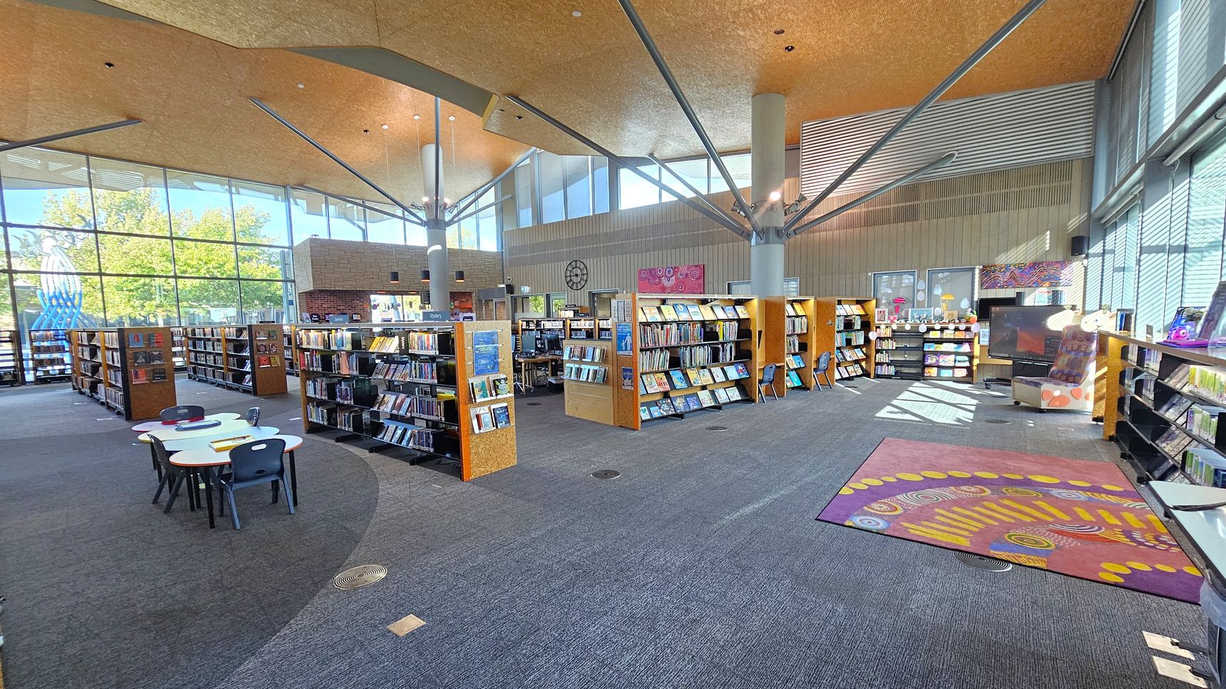 A spacious library interior with large windows on the left, allowing natural light to illuminate the space. In the foreground, there are small white tables with black chairs. The middle area features rows of wooden bookshelves filled with books. On the right, a colourful rug with circular patterns is placed on the grey carpeted floor. The ceiling is high with exposed beams and a warm, wooden finish. A large TV and more bookshelves are visible in the background.