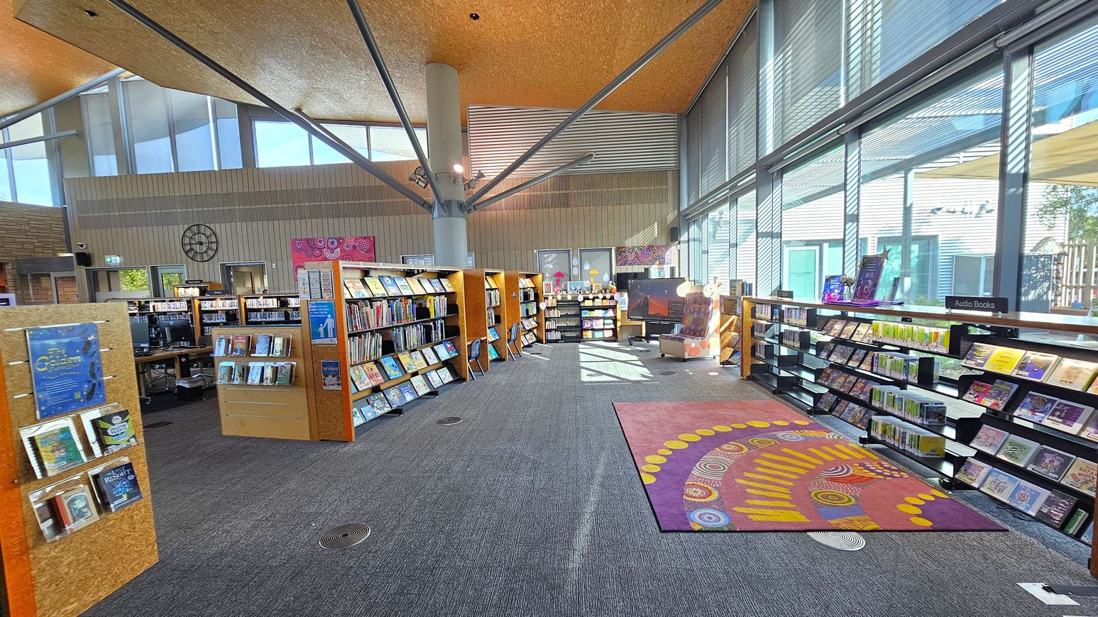 A library interior with a modern design. On the left, there are bookshelves filled with books and a display stand with posters. In the centre, more bookshelves are arranged in a semi-circle, with a large pillar supporting the ceiling. On the right, a long shelf holds audio books, and large windows let in natural light. The floor is carpeted with a colourful patterned rug in the middle. The ceiling is high with exposed beams, and there are artworks and a clock on the walls.