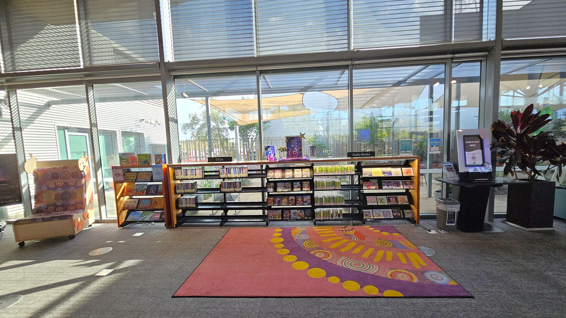 A library interior with large glass windows in the background, allowing natural light to illuminate the space. In the centre, there are shelves filled with DVDs and audiobooks. To the left, a colourful chair with a patterned design. On the right, a digital check-out kiosk next to a potted plant. The floor is carpeted with a vibrant rug featuring circular patterns in the foreground.