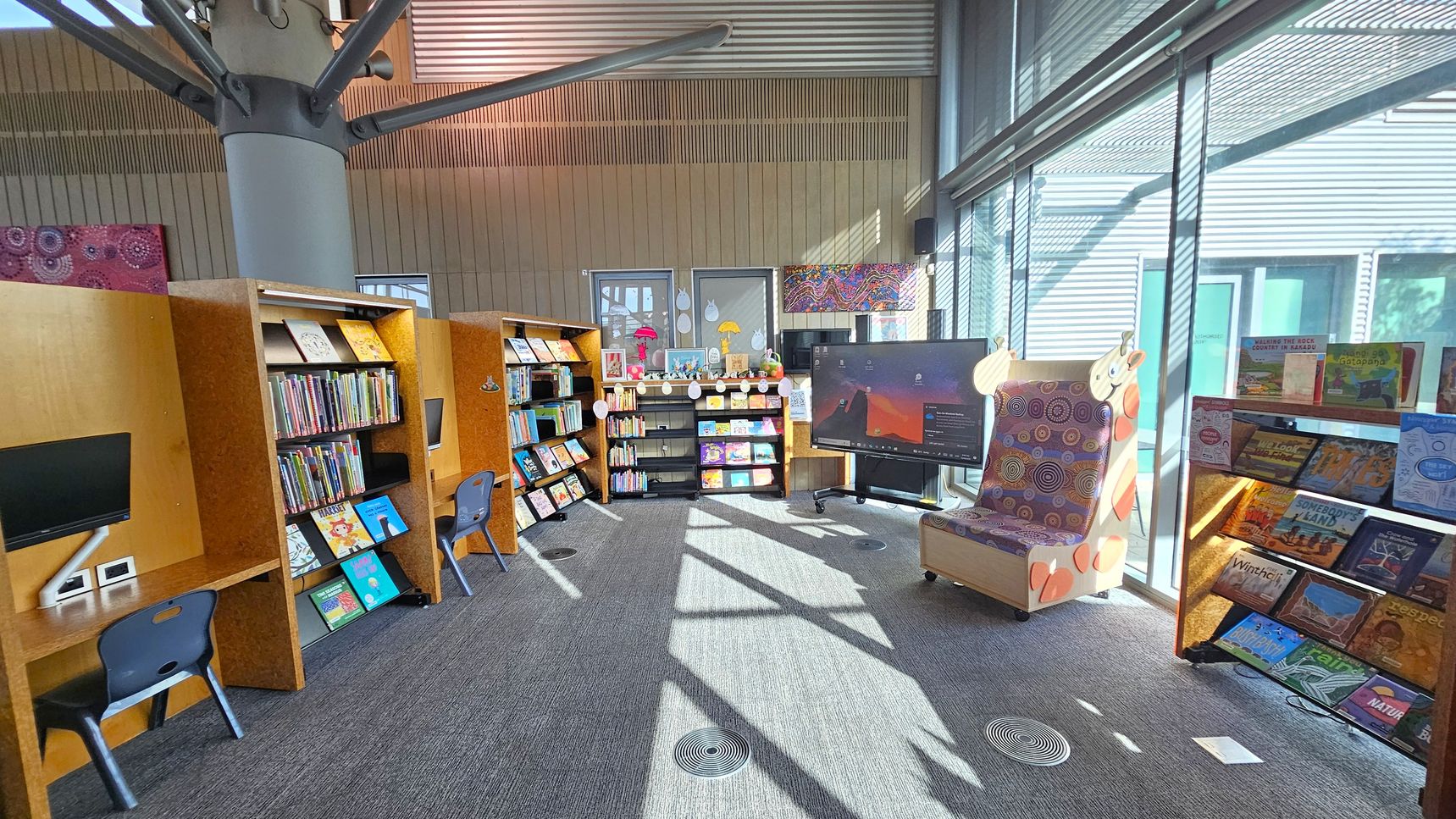A library space with bookshelves filled with children's books on the left and centre. Two computer stations with blue chairs are on the left. In the centre, a small black bookshelf holds more books and decorations. A large digital screen is on the right, next to a colourful giraffe-themed chair. The right side features a window with blinds, allowing sunlight to illuminate the room. The floor is carpeted, and the ceiling has exposed beams and a large column.