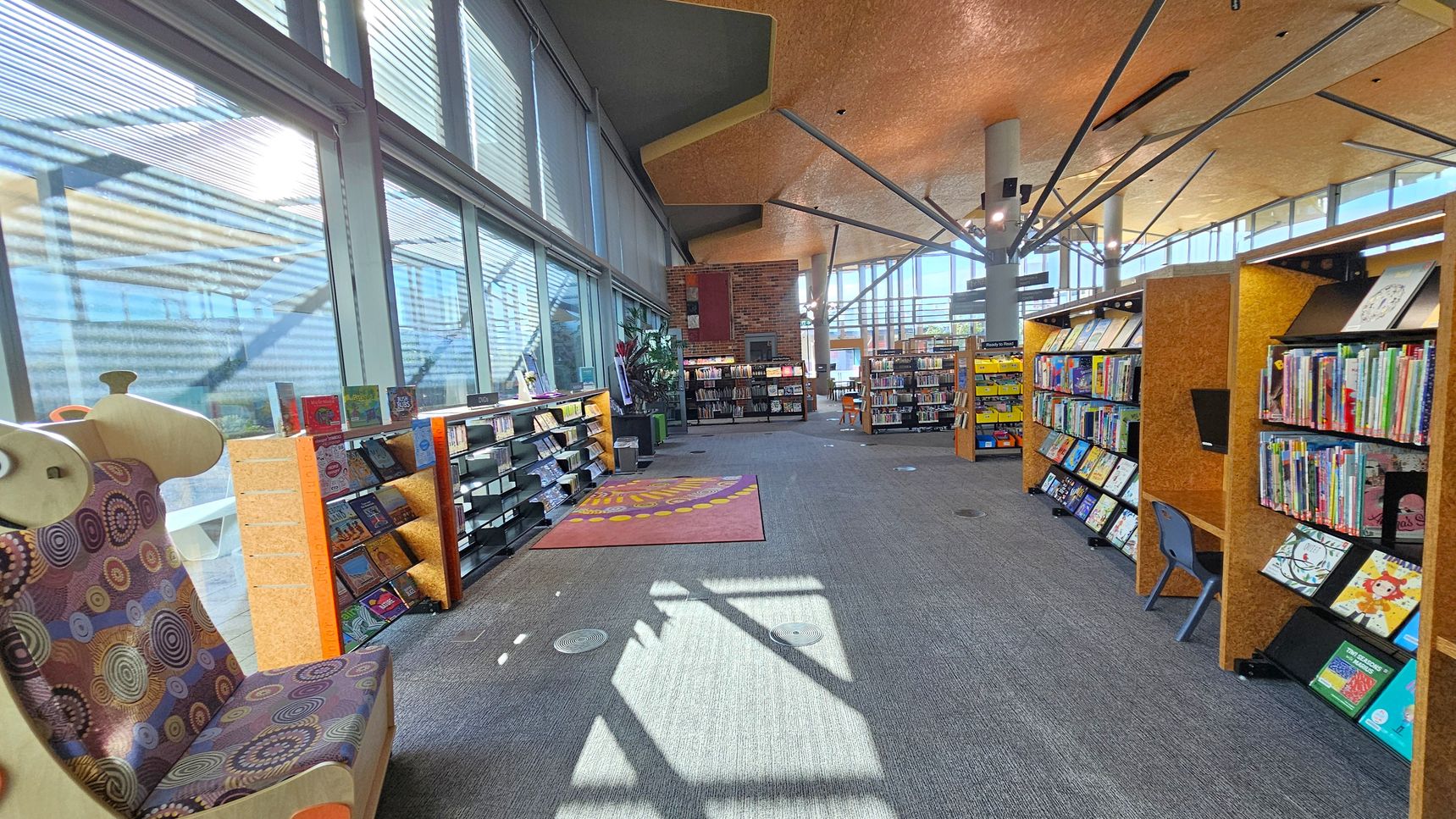 A library interior with large windows on the left, allowing natural light to stream in. A patterned chair is near the windows, and bookshelves line the walls, filled with colourful books. In the centre, a carpet with a circular design lies on the grey carpeted floor. The ceiling is high with exposed beams. In the background, more bookshelves and a small table with chairs are visible, creating a spacious and inviting reading area.