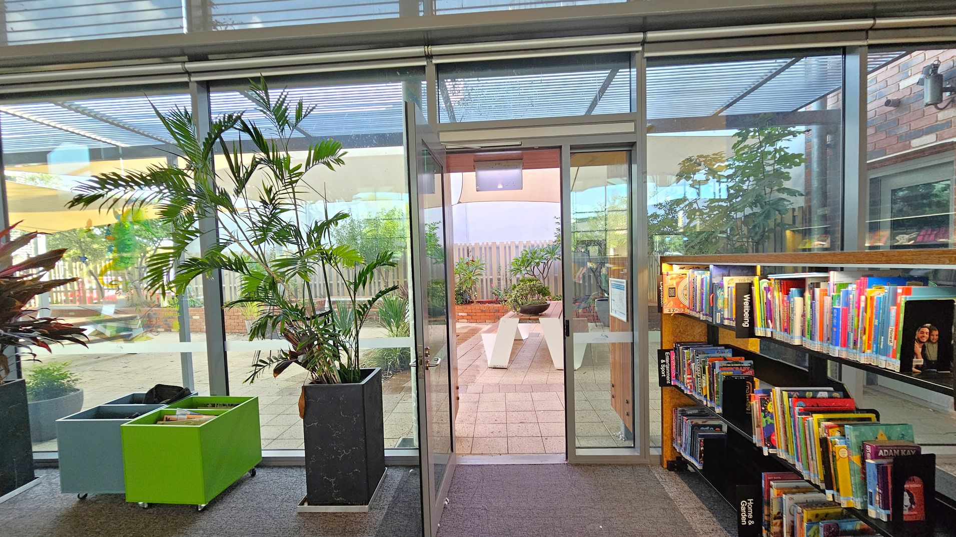 A library interior with a glass door in the centre leading to an outdoor area with plants and benches. On the left, there are two mobile book bins, one green and one blue, next to a tall potted plant. On the right, a bookshelf filled with colourful books lines the wall. The floor is carpeted, and the ceiling has a grid-like structure with glass panels.