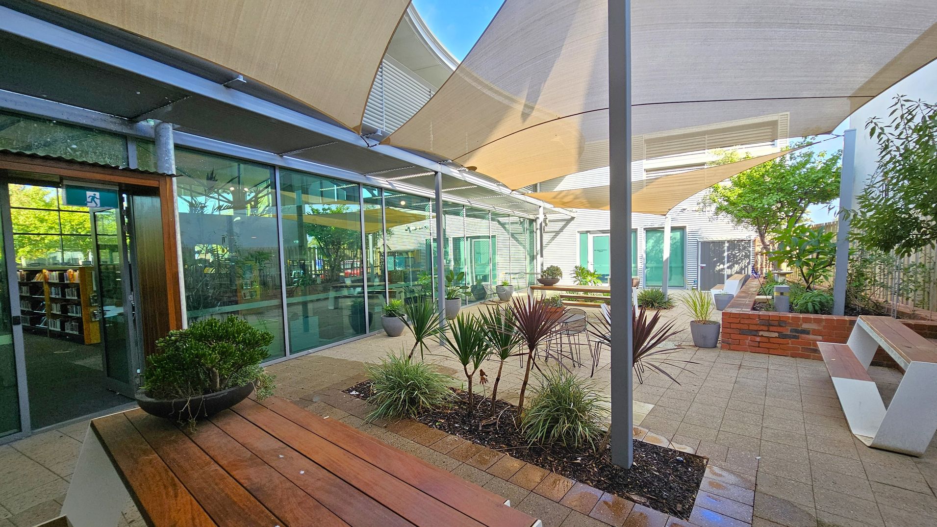 A courtyard with a paved floor and several potted plants. On the left, there's a glass wall with a view into a room containing bookshelves. A wooden bench with a plant on it is in the foreground. Overhead, there are large fabric canopies providing shade. On the right, a brick planter with more greenery and a white bench is visible. The background features a building with green doors and windows, and more plants are scattered throughout the area.