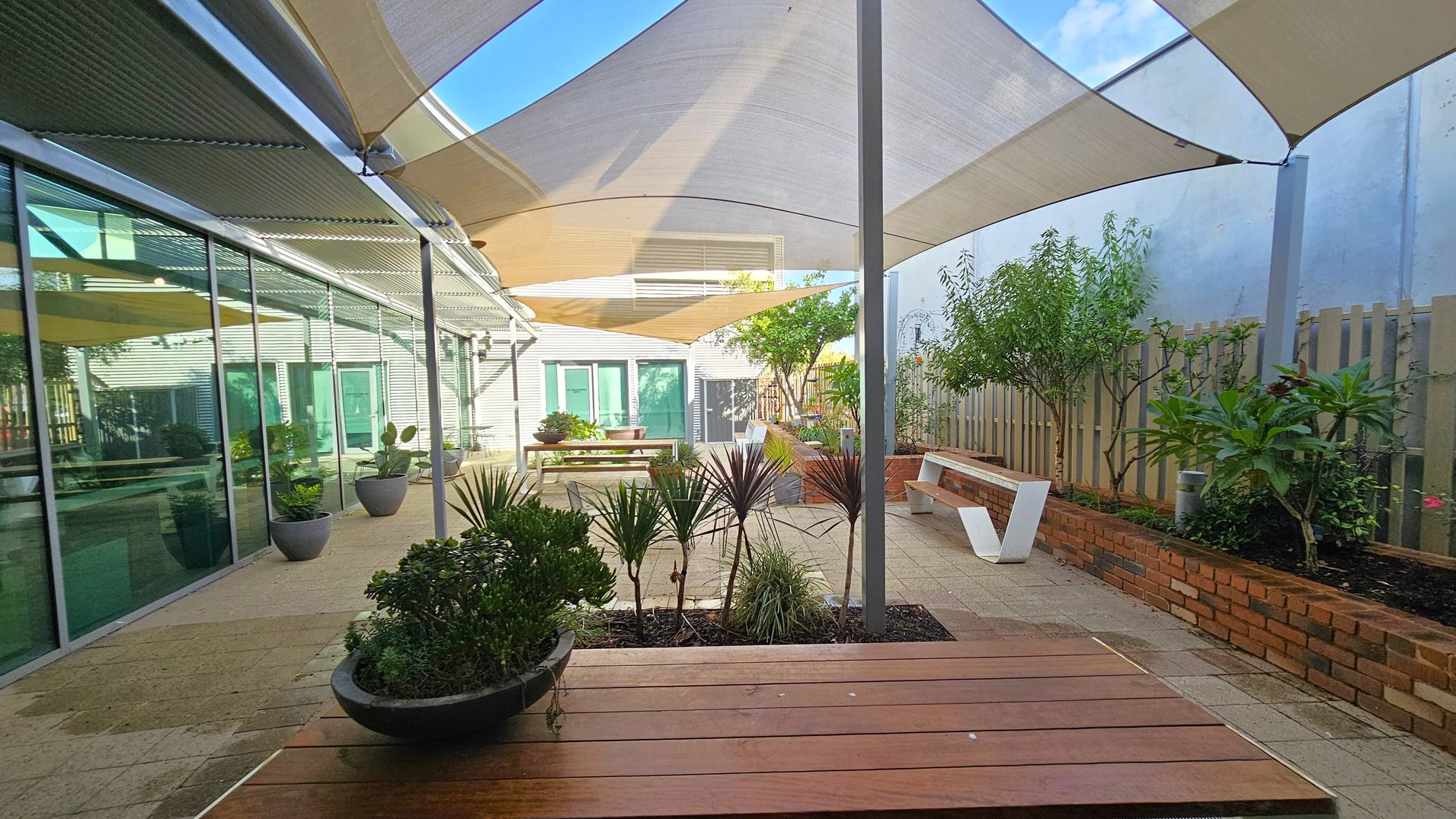 A courtyard with a paved floor and several triangular shade sails overhead. On the left, there is a glass wall with potted plants in front. In the centre, a wooden bench and a planter with various plants are visible. On the right, a brick planter box with small trees and shrubs lines the area, accompanied by a modern white bench. A wooden fence runs along the right side, and the background features a building with green doors and windows.