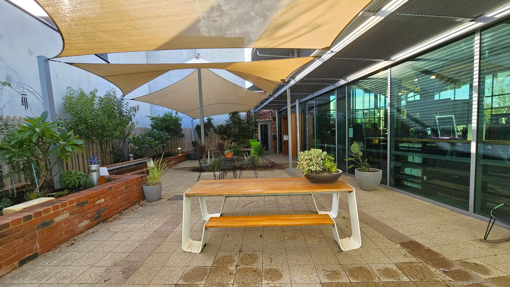 A courtyard with a wooden table and bench in the foreground, topped with a potted plant. To the right, a glass wall reflects the interior of a building. The left side features a brick planter with various green plants and a wooden fence. Overhead, beige triangular shade sails provide cover. The floor is paved with light-coloured tiles, and there are additional potted plants and outdoor seating in the background.