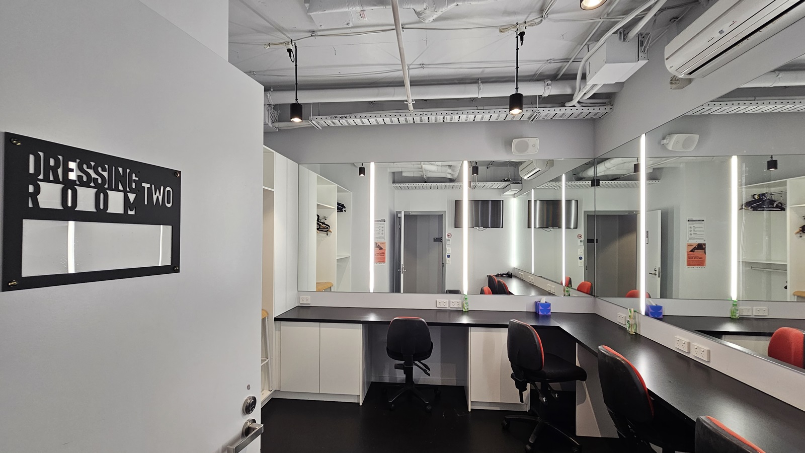 Dressing Room Two has mirrors fixed to the wall and adjustable chairs nearby. An inward-opening door with a sign is to the left. The ceiling shows exposed concrete beams, pipes, and air conditioning vents. The walls are white, and the floor is tiled black.
