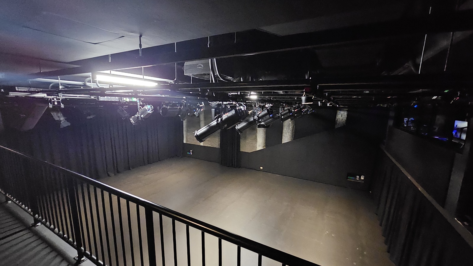 A dimly lit space with black floor tiles and walls. Metal railing secures the area. Overlooking below is the stage with black tiles, surrounded by black walls and curtains. The ceiling features exposed pipes, audio equipment, and lighting fixtures.