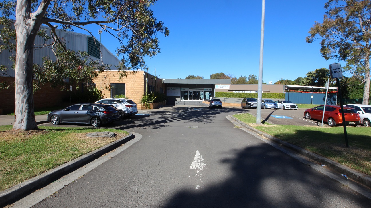 The view metres away from the front of Michael Wendon Aquatic Leisure Centre. Michael Wenden ALC is a low-set brown and grey industrial, warehouse-style building. The accessible car parks available in front of the venue are marked by an accessible parking sign painted in white and blue. On the right side of the venue is a ramped access that allows for a step-free entrance with four grey steps to enter the venue through automatic glass sliding doors. The view metres away from the front of Michael Wendon Aquatic Leisure Centre. Michael Wenden ALC is a low-set brown and grey industrial, warehouse-style building. The accessible car parks available in front of the venue are marked by an accessible parking sign painted in white and blue. On the right side of the venue is a ramped access that allows for a step-free entrance with four grey steps to enter the venue through automatic glass sliding doors.