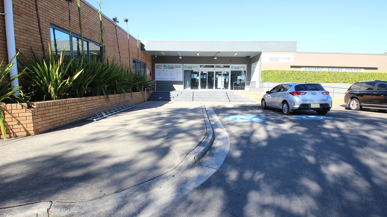Concrete pathway leading to the four-step grey stairs with three metal handrails to the entrance of the Michael Wendon Aquatic Leisure Centre. Accessible car parking spaces can be found close to and directly opposite the entrance doors. These doors are lined with white edges. In this image, the plants with long leaves in the garden to the left covered the glass windows and had a long stem at the center leveling the topmost part of the wall. Concrete pathway leading to the four-step grey stairs with three metal handrails to the entrance of the Michael Wendon Aquatic Leisure Centre. Accessible car parking spaces can be found close to and directly opposite the entrance doors. These doors are lined with white edges. In this image, the plants with long leaves in the garden to the left covered the glass windows and had a long stem at the center leveling the topmost part of the wall.