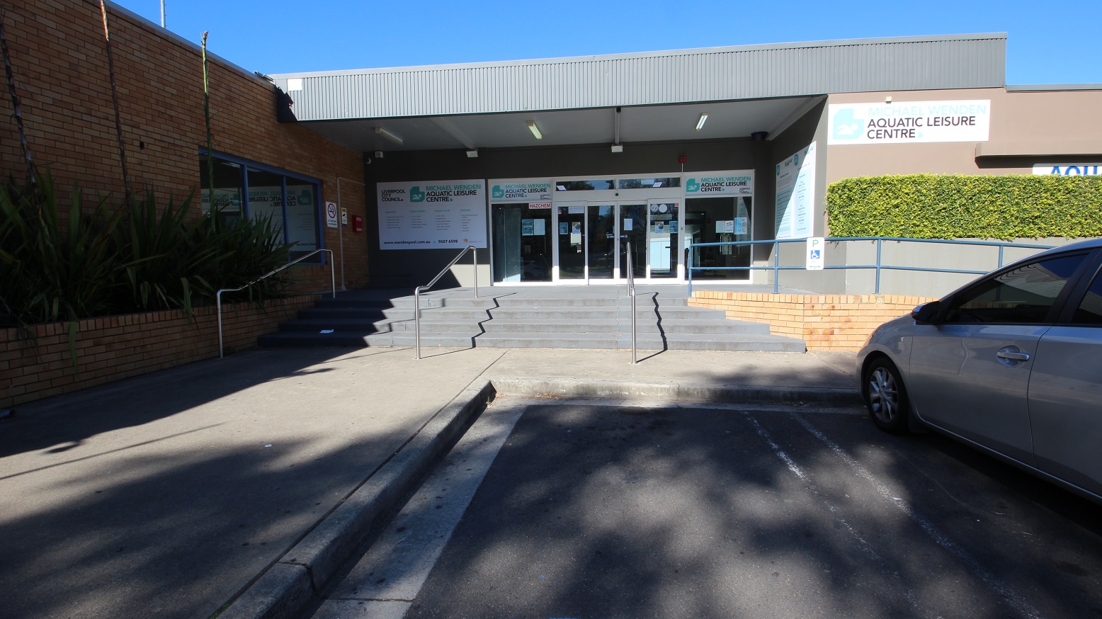 On the blue handrail near the automatic glass sliding doors is the accessible parking sign in white and blue. Bushes cleanly trimmed filled the garden against the smooth brown wall on the right side of the Michael Wendon Aquatic Leisure Centre. Several marketing and informative posters in different sizes are placed on the walls of the entrance doors. There is also a no-smoking sign, and a CCTV camera in operation can be seen on the brick wall to the left of the entrance wall. On the blue handrail near the automatic glass sliding doors is the accessible parking sign in white and blue. Bushes cleanly trimmed filled the garden against the smooth brown wall on the right side of the Michael Wendon Aquatic Leisure Centre. Several marketing and informative posters in different sizes are placed on the walls of the entrance doors. There is also a no-smoking sign, and a CCTV camera in operation can be seen on the brick wall to the left of the entrance wall.