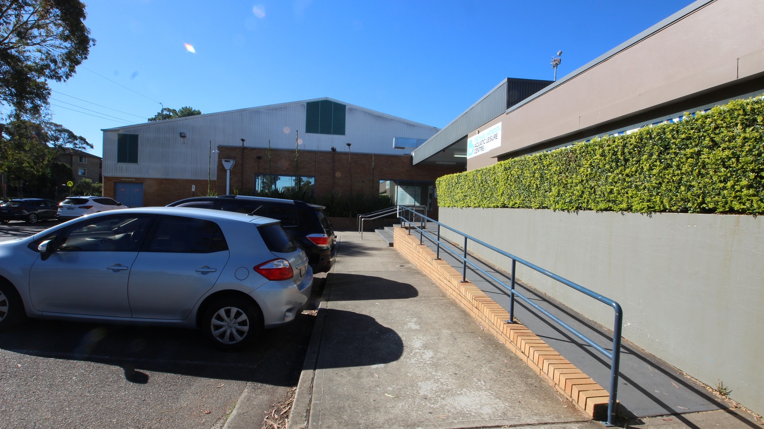Image shows a concrete pathway before the grey stairs and the accessible ramped access at Michael Wendon Aquatic Leisure Centre. There are tall trees with significant foliage that provide shade to the cars at the parking spaces close to the entrance. From this side, a part of the venue with a white-painted wall and lines can be seen behind the brick wall. It has two windows for air ventilation. Image shows a concrete pathway before the grey stairs and the accessible ramped access at Michael Wendon Aquatic Leisure Centre. There are tall trees with significant foliage that provide shade to the cars at the parking spaces close to the entrance. From this side, a part of the venue with a white-painted wall and lines can be seen behind the brick wall. It has two windows for air ventilation.