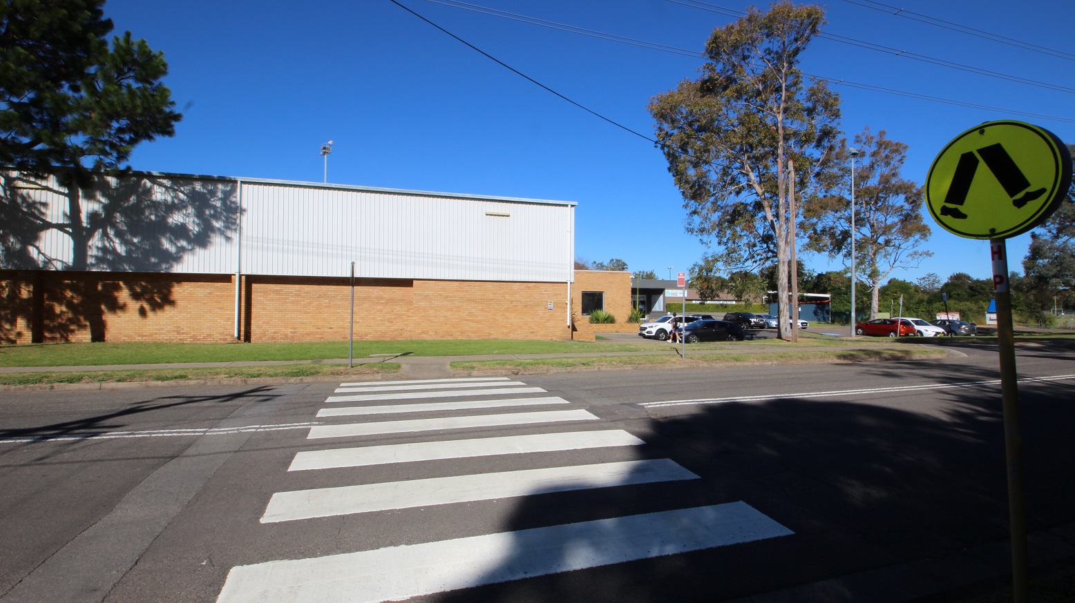 Pedestrian lane leading to the left side of the Michael Wendon Aquatic Leisure Centre. It can be identified by the neon green pedestrian crossing sign containing a walking leg icon and shoes. The lane can be identified by the white Zebra lanes. Cars of various makes and colors are parked in the area in front of the venue. The parking spots closest to the entrance are shaded by tall trees with ample foliage. Pedestrian lane leading to the left side of the Michael Wendon Aquatic Leisure Centre. It can be identified by the neon green pedestrian crossing sign containing a walking leg icon and shoes. The lane can be identified by the white Zebra lanes. Cars of various makes and colors are parked in the area in front of the venue. The parking spots closest to the entrance are shaded by tall trees with ample foliage.